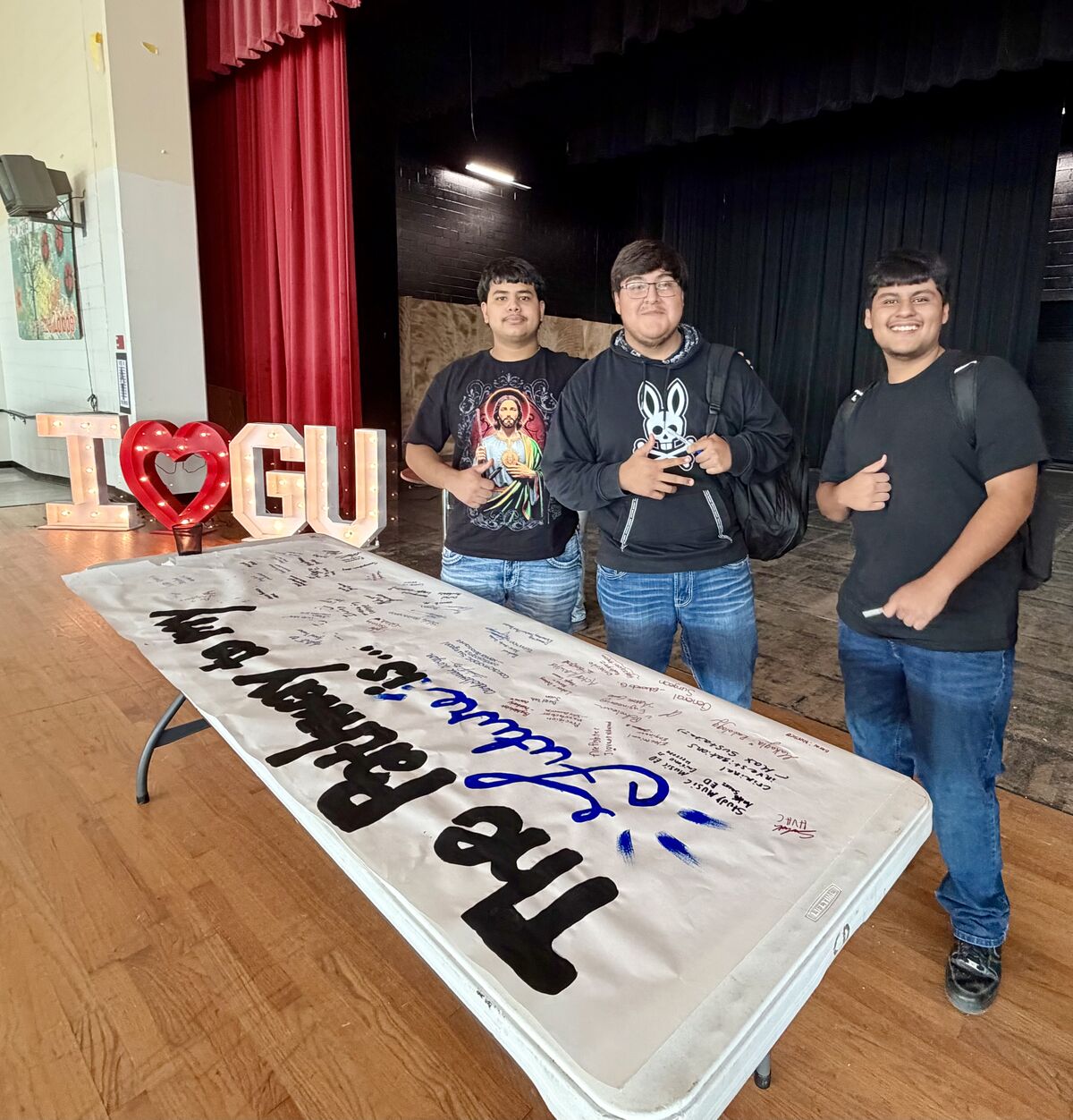 Three smiling students stand behind a signed banner on a table, with illuminated "I HEART U" letters in the background.