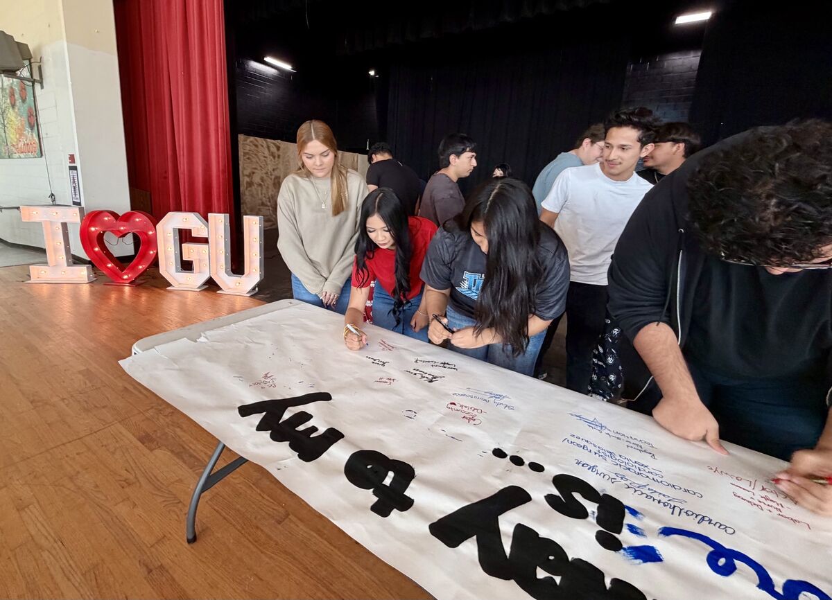 A group of students gather around a long table, signing a large paper banner in an indoor hall.