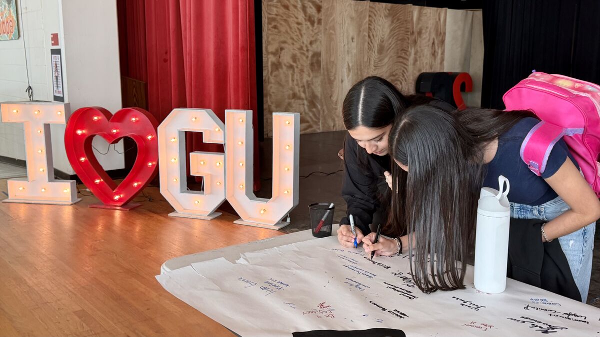 Two people lean over a long white paper laid on a table, writing signatures with markers.
