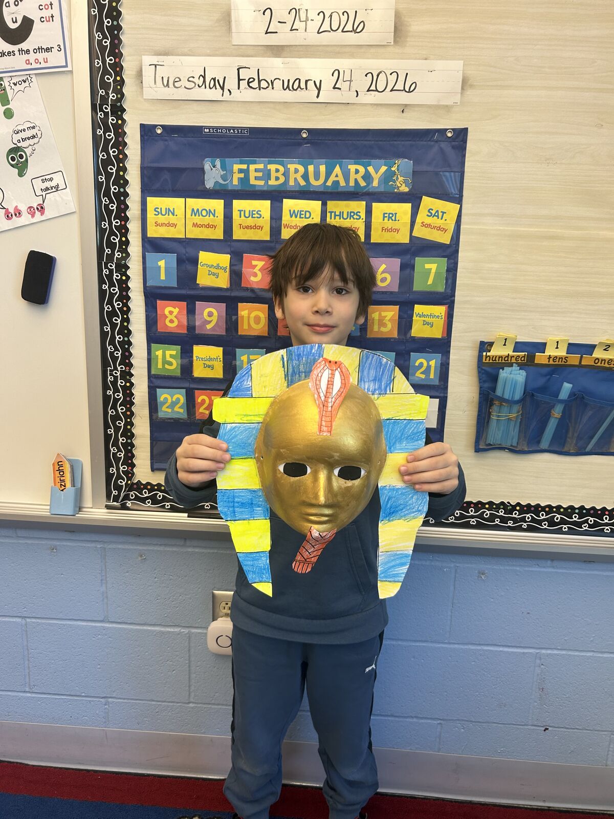 A child holds up a handmade Egyptian pharaoh mask in front of a February calendar.