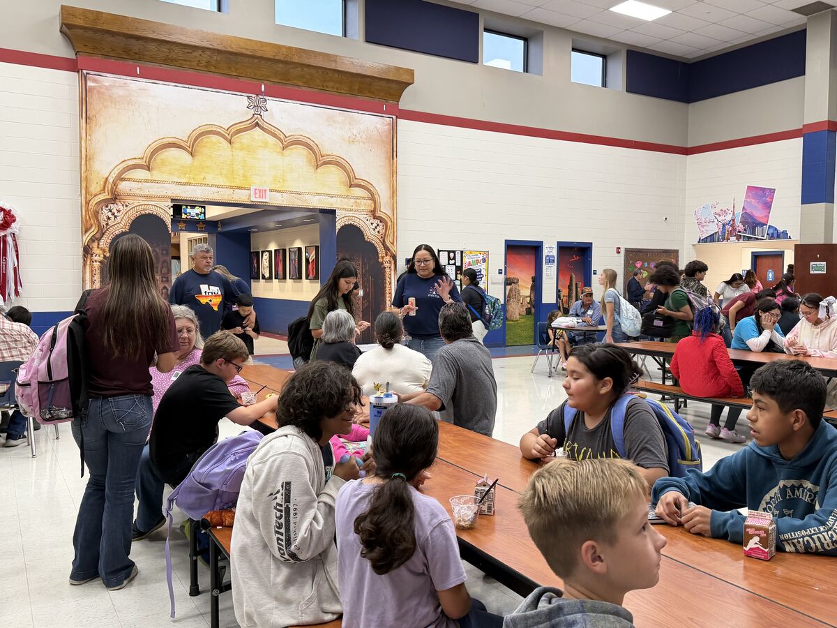 Students and adults gather in a brightly lit school cafeteria with a large mural of an Indian archway on the back wall.