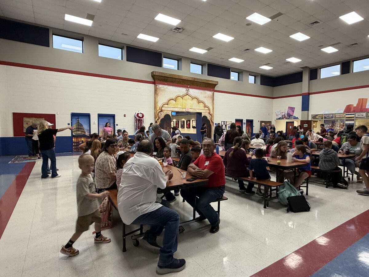 A wide-angle view of a large, brightly lit cafeteria filled with people sitting at long tables, eating and talking.