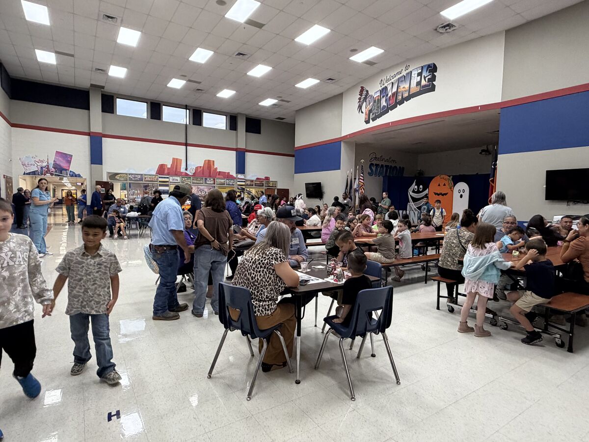 A lively school cafeteria filled with children and adults sitting at tables and walking around, with a large 'Welcome to Pawnee' sign on the wall.