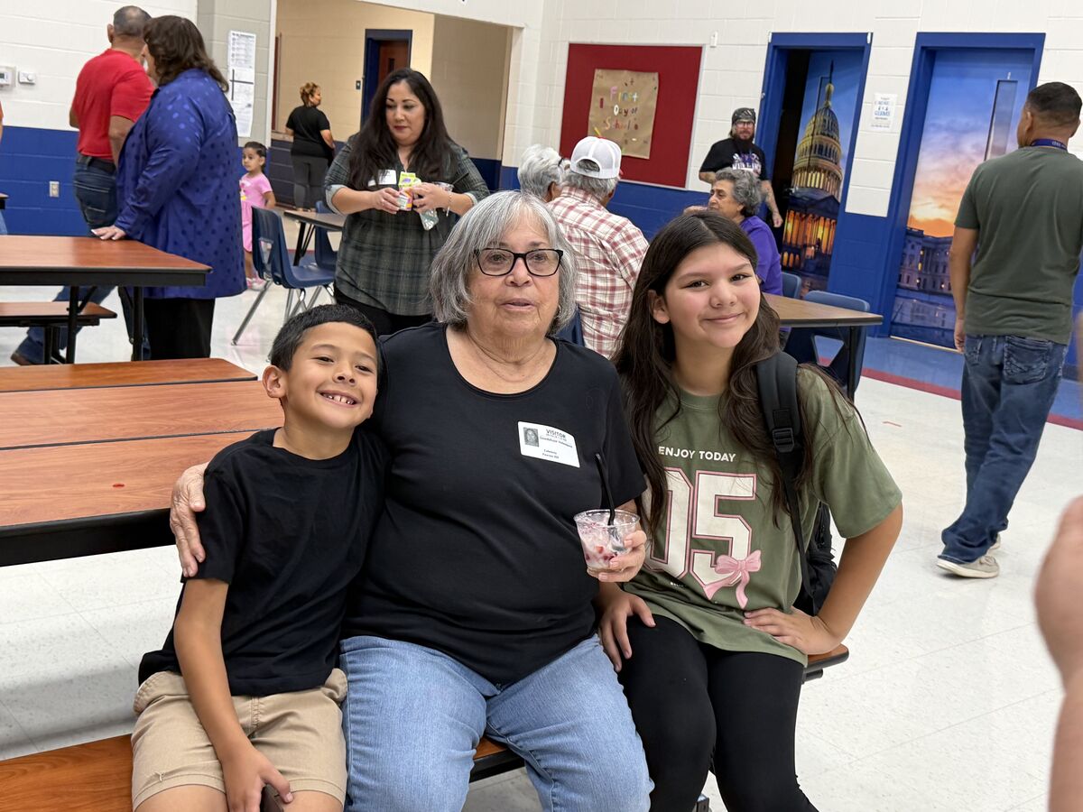 A grandmother sits between her smiling grandchildren in a school cafeteria.