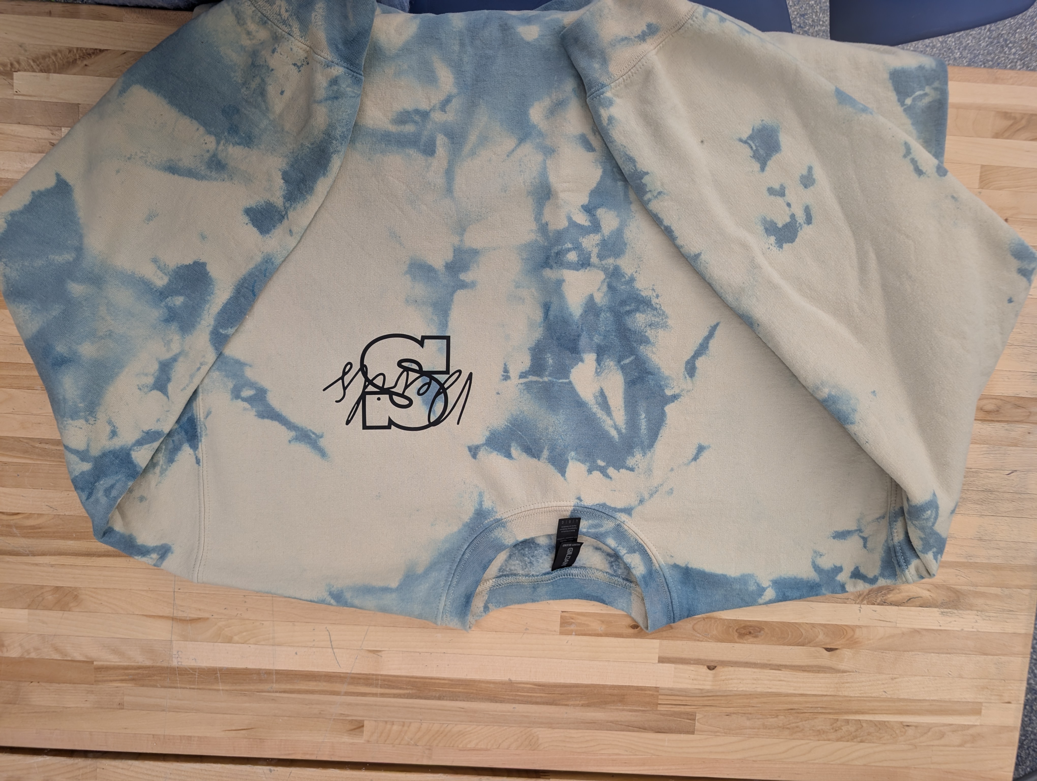 A light-colored sweatshirt with a blue tie-dye pattern and a black graphic logo.