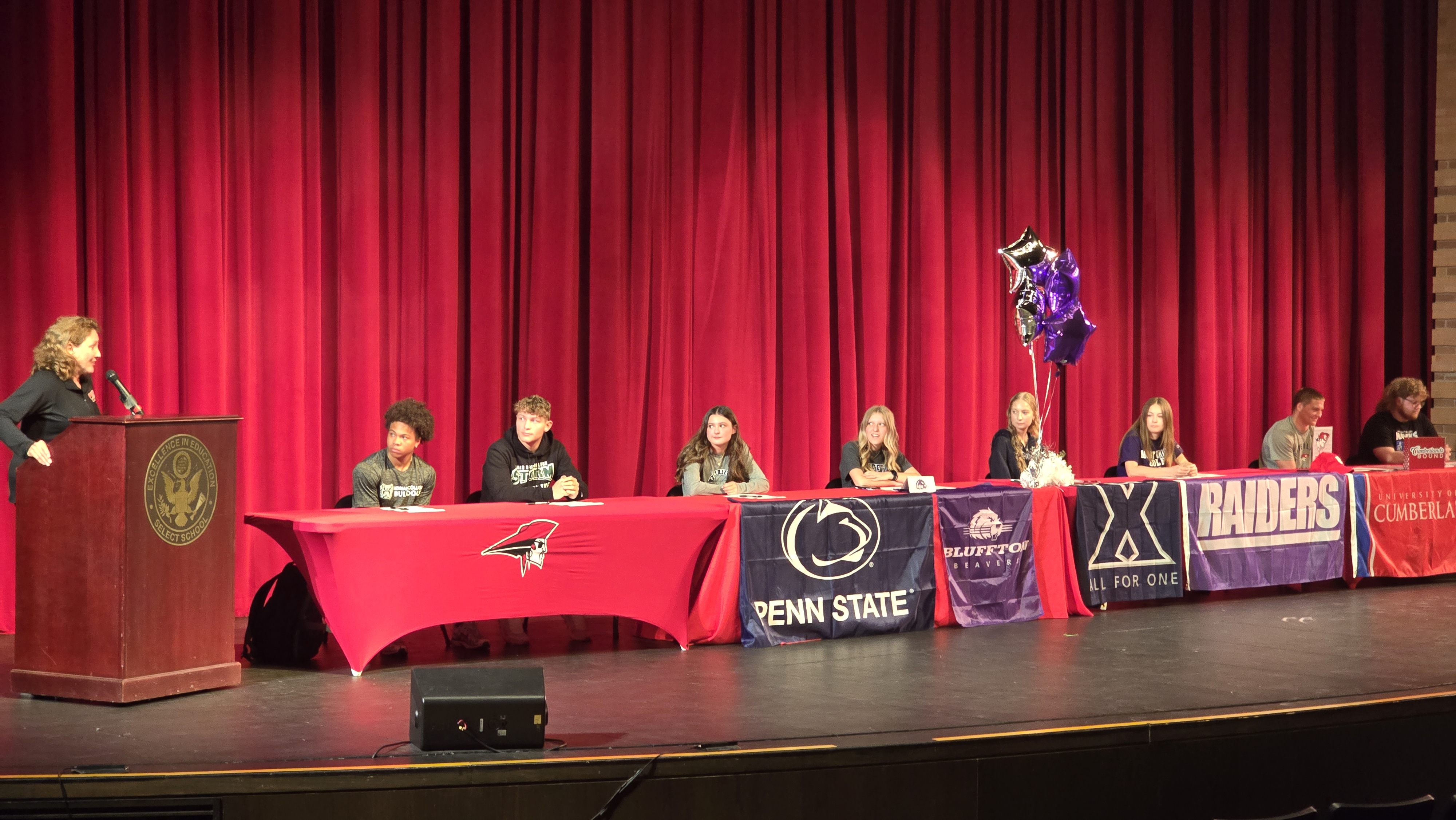 A group of students sit at a table on a stage in front of a red curtain, with banners representing different universities.