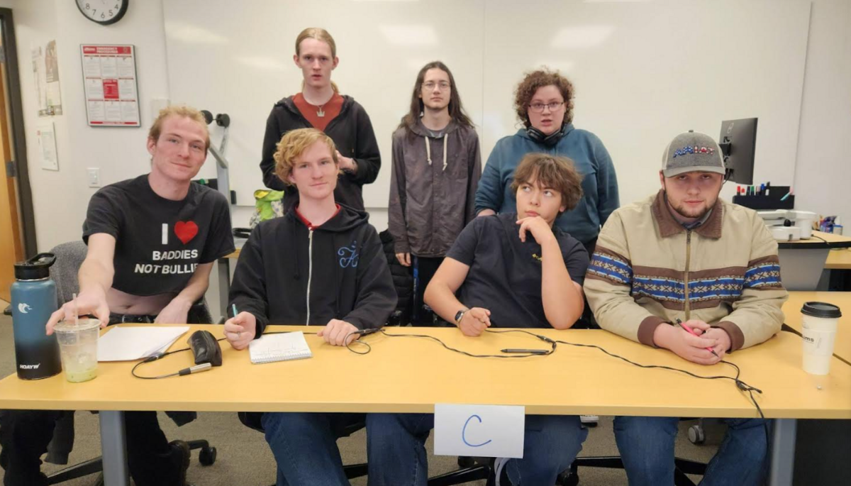 Seven young people pose for a photo behind a long table in a classroom setting.