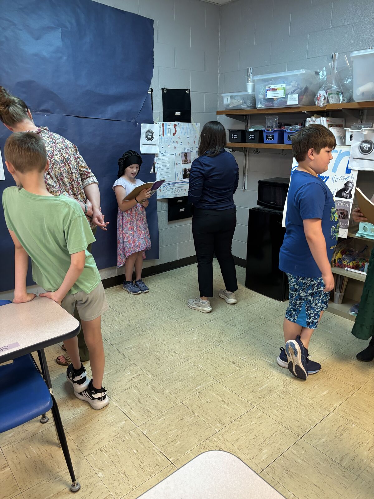 Students gather in a classroom, some standing and others sitting on the floor, with educational posters on the wall.