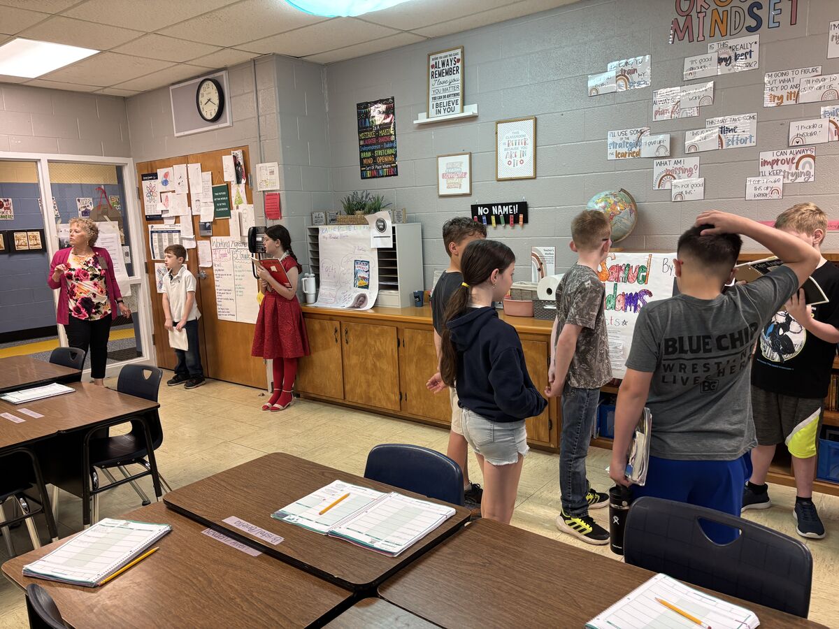 A teacher stands in a classroom, observing students gathered around a display board with various posters and student work.