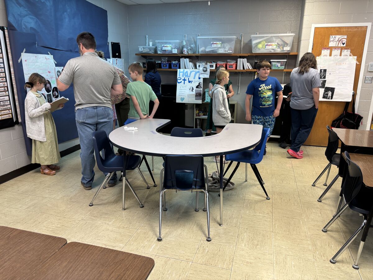 Students and adults gather around a horseshoe-shaped table in a classroom, looking at displays on the wall.
