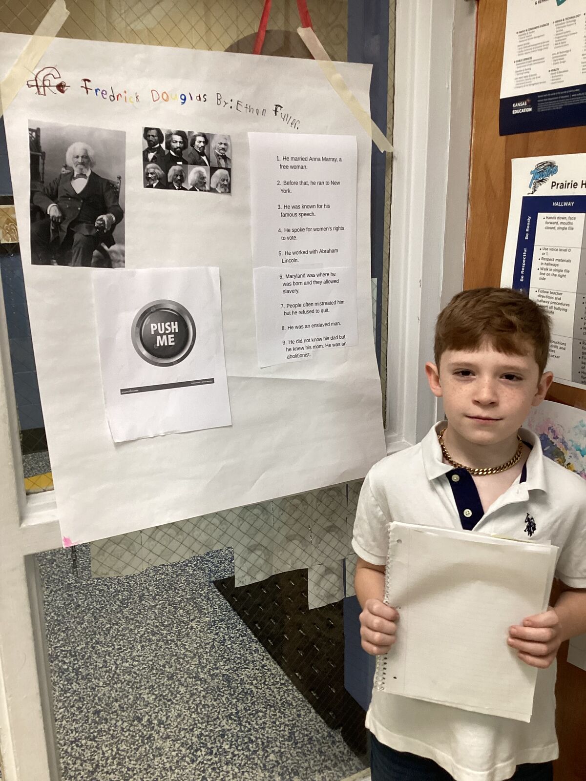 A young boy stands next to a school project about Frederick Douglass, featuring historical photos and biographical facts.