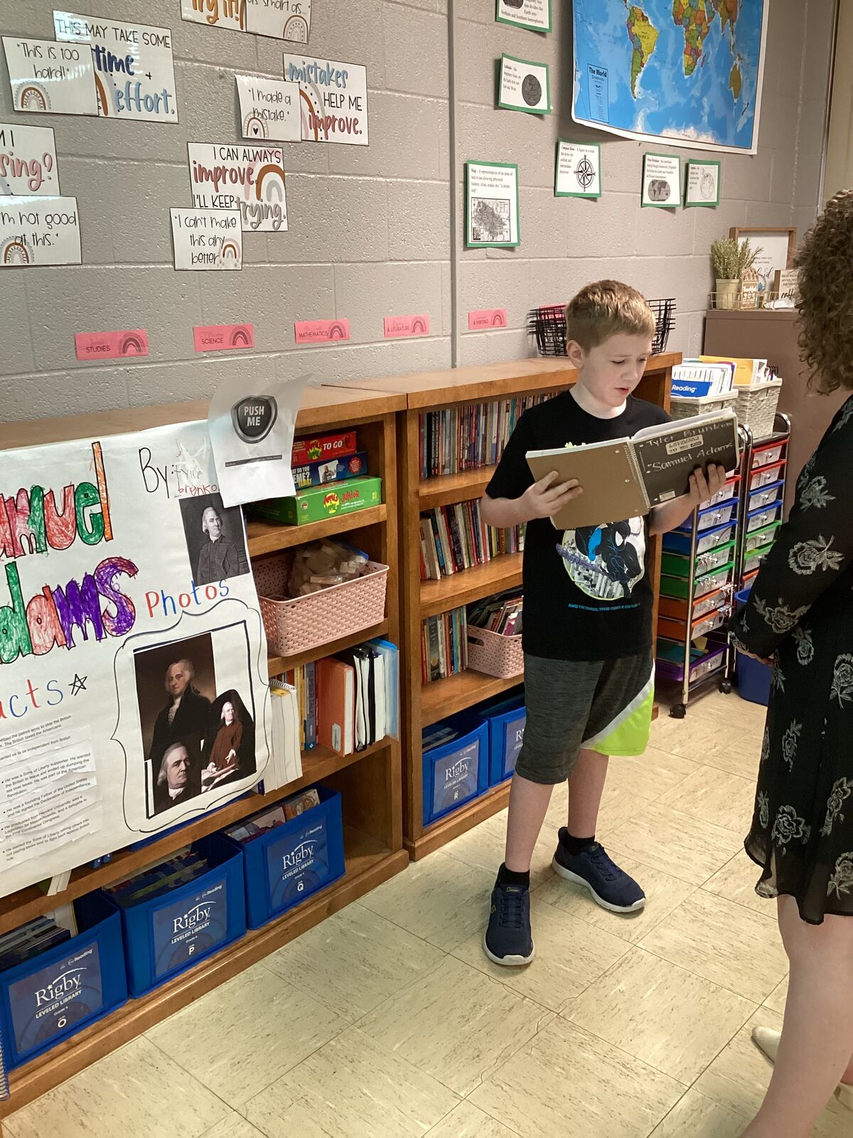 A young boy stands in a classroom, holding a notebook and looking at a display board with historical photos and text.