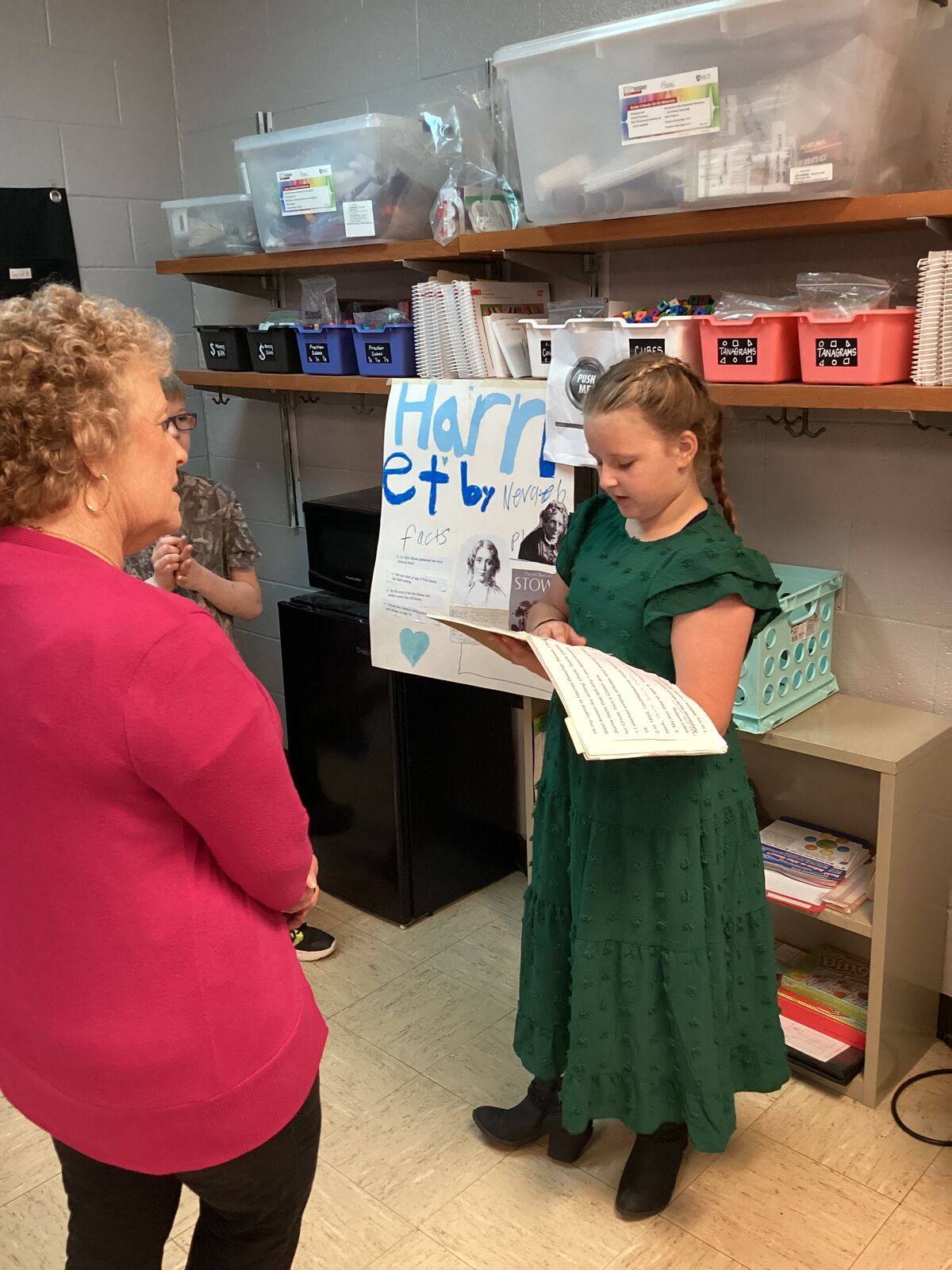 A young person in a green dress presents a poster about Harriet Beecher Stowe to an adult.
