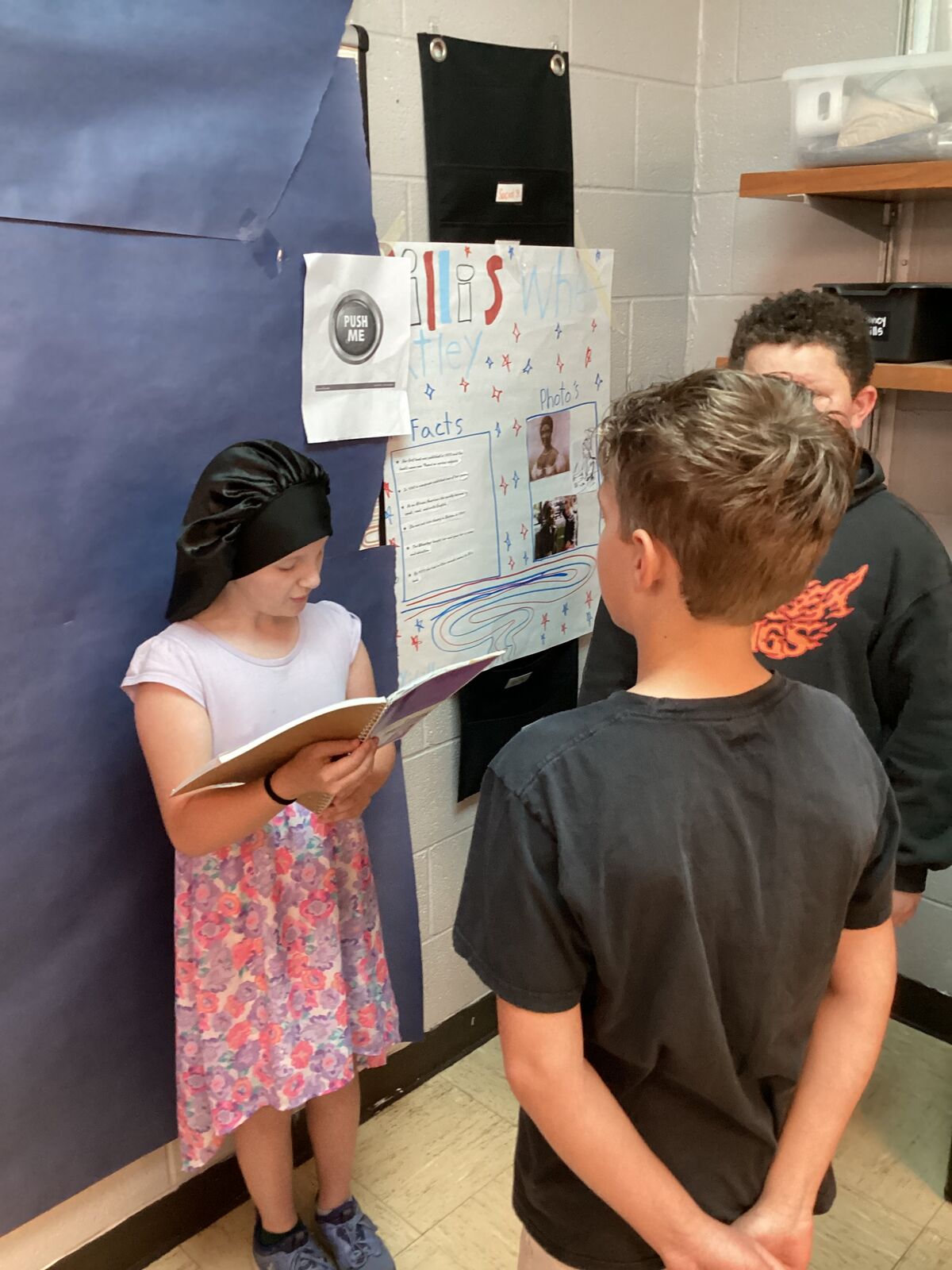 Two students look at a project board with photos and facts, while another student reads a notebook.