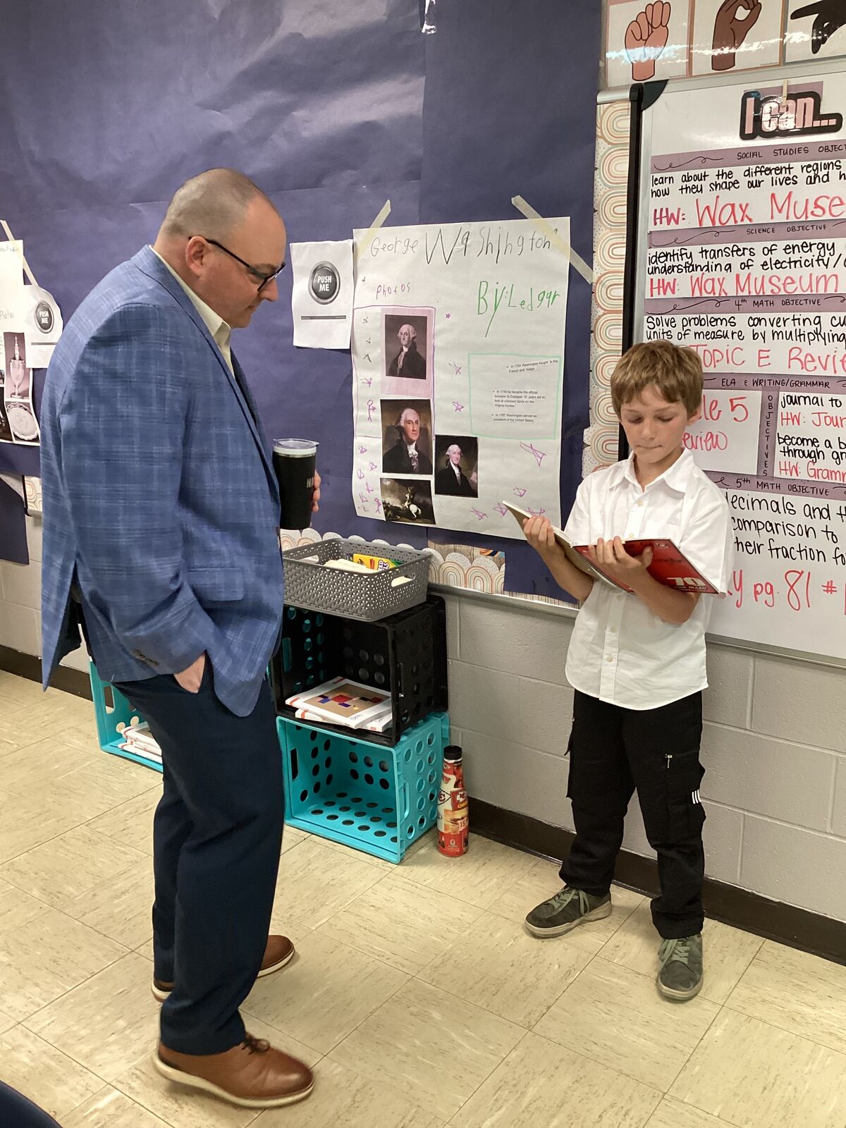 The superintendent in a blue suit looks up at a student holding a book in a classroom.