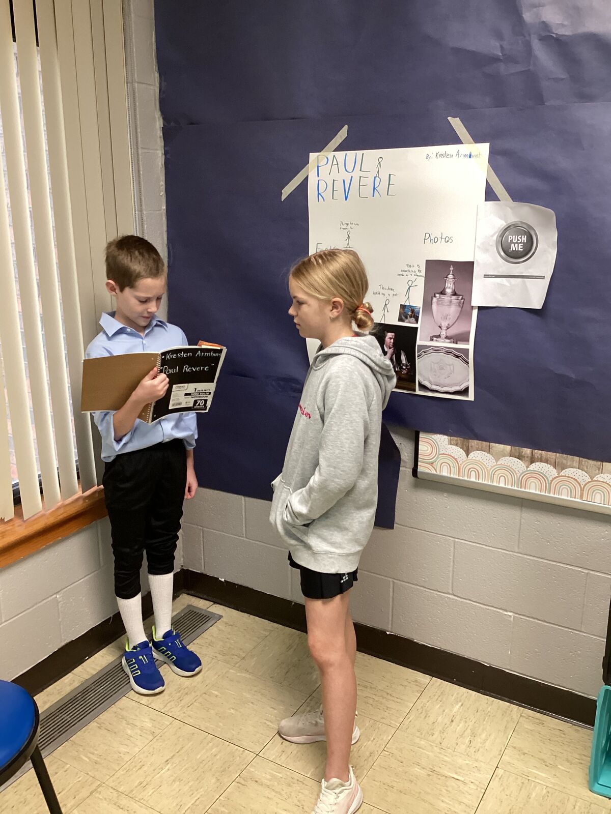 Two students stand near a bulletin board displaying information about Paul Revere, with one holding a notebook.