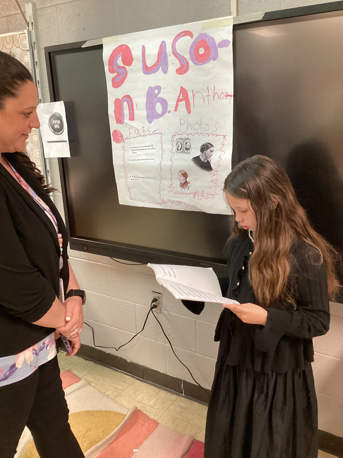 A young girl in a black dress reads from a paper in front of a large screen and a colorful poster.