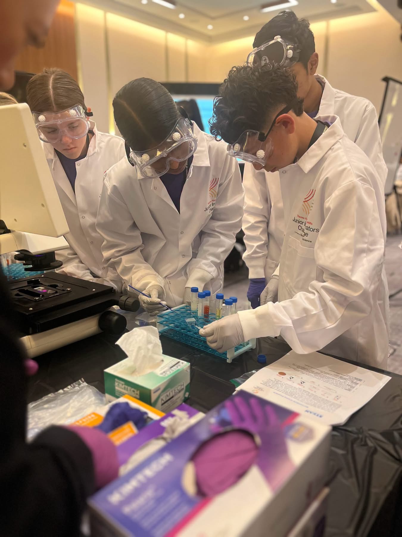 Four students wearing lab coats and safety goggles conduct a science experiment in a bright room.