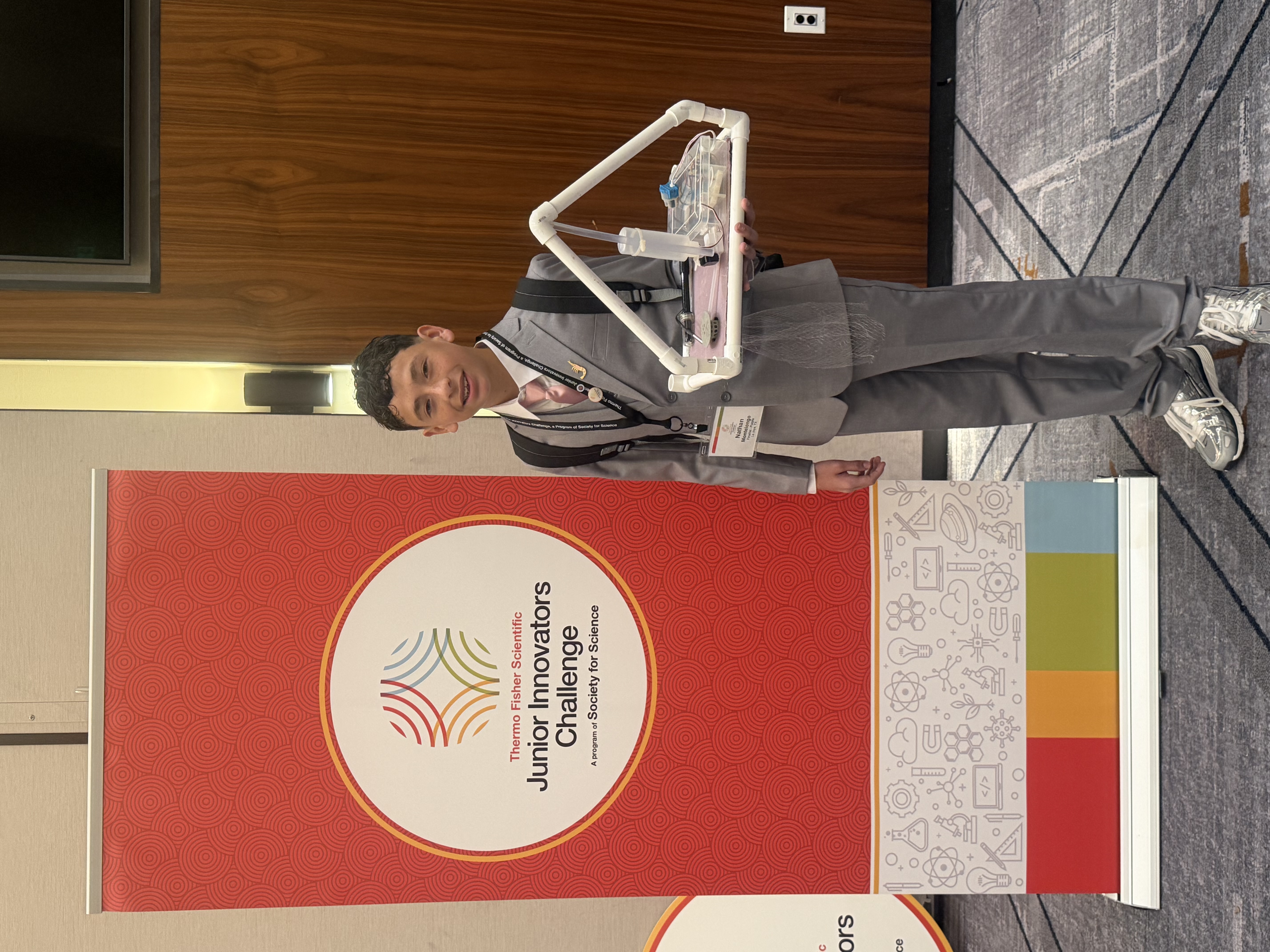 A young person in a grey suit smiles while holding a science project next to a Thermo Fisher Scientific Junior Innovators Challenge banner.
