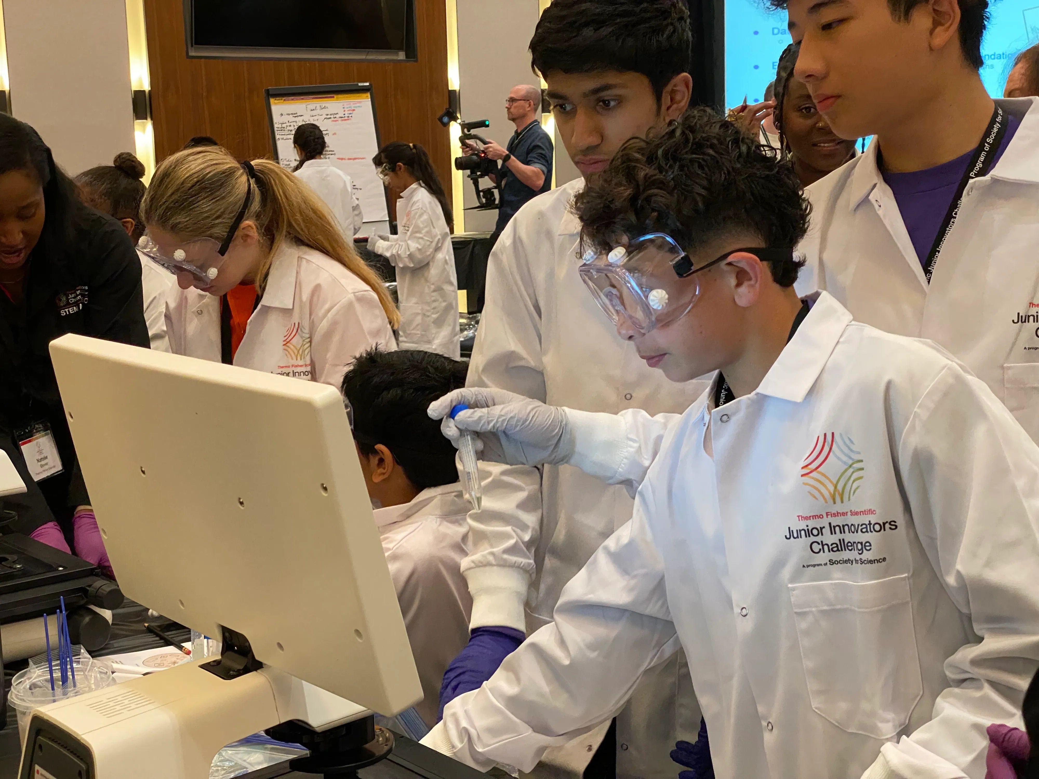 Students in white lab coats and safety goggles conduct experiments and examine equipment.