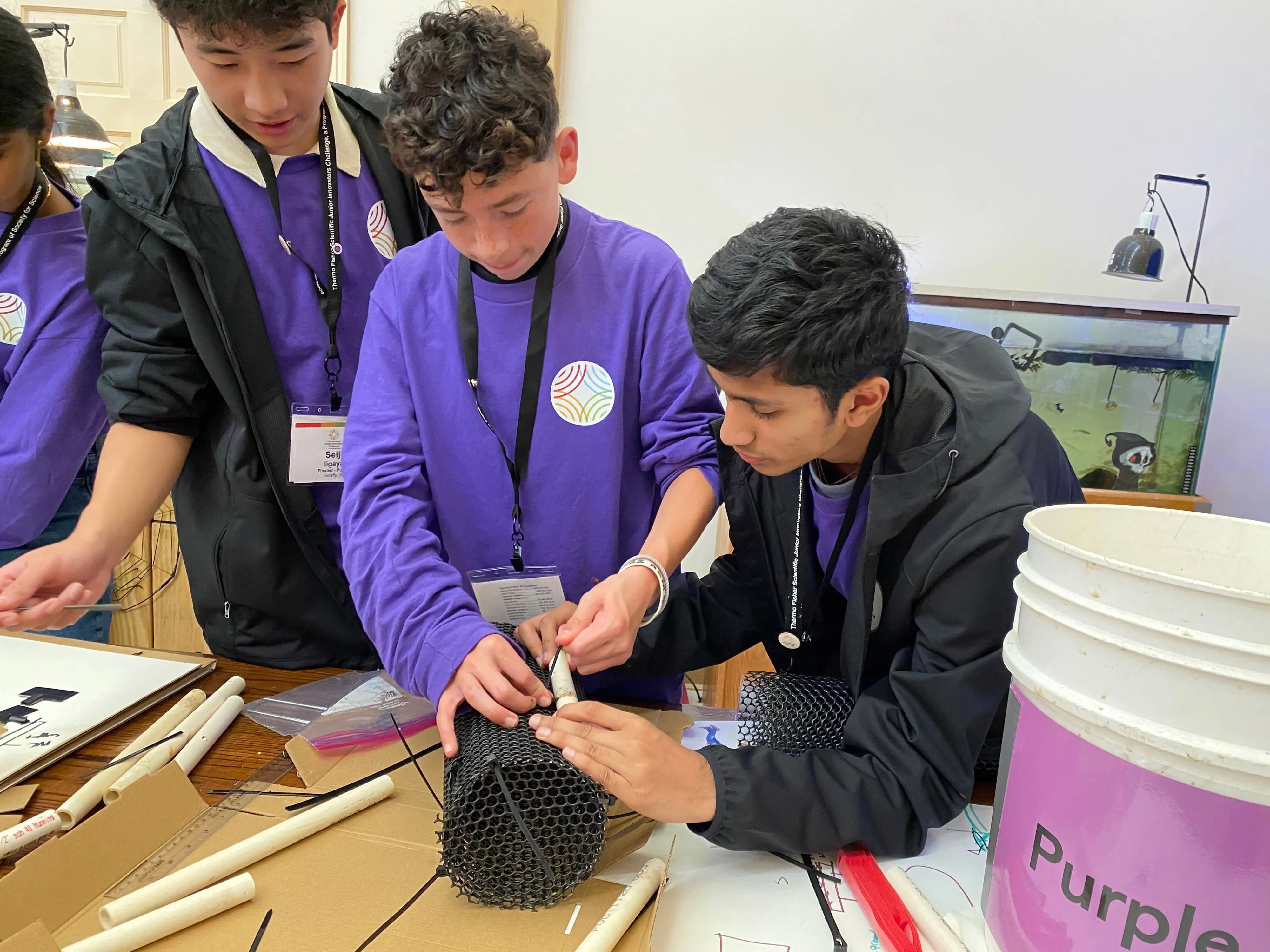 Three young people wearing matching purple shirts work together at a table to assemble a mesh and plastic project.
