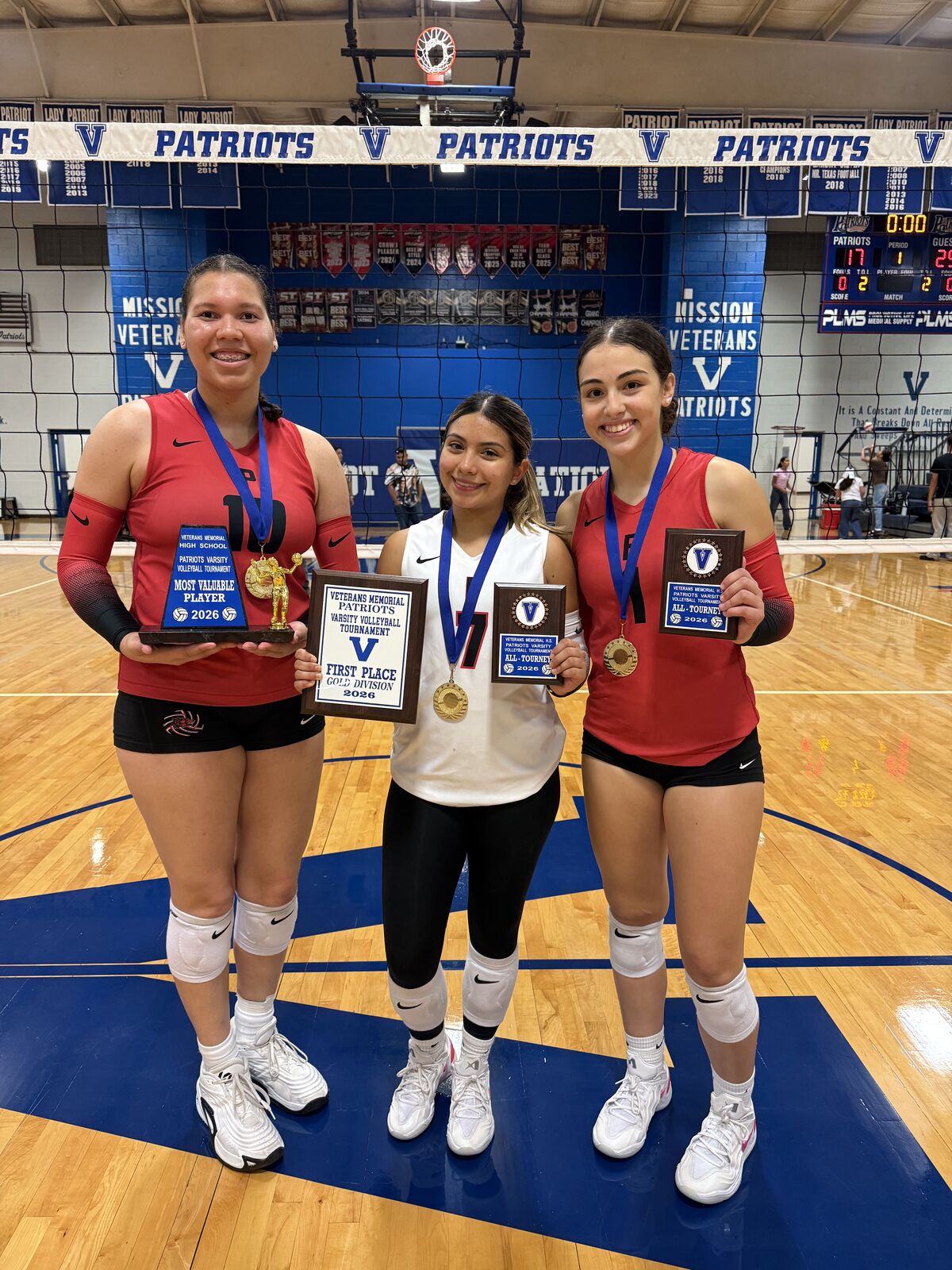 Three smiling young women in volleyball uniforms hold trophies and medals on a gymnasium court.