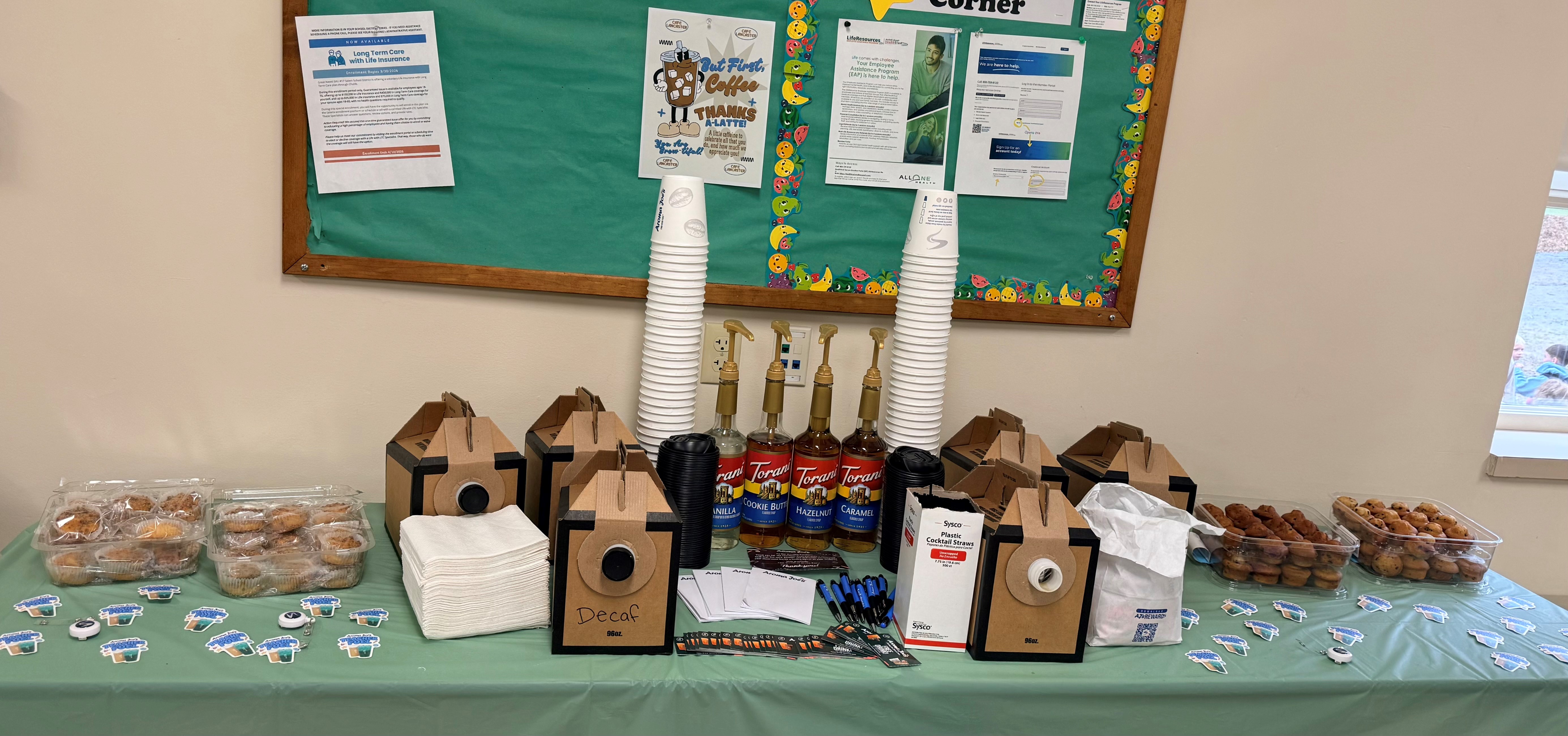 A coffee and pastry station is set up on a table with a green tablecloth, featuring Torani syrups, coffee boxes, and stacks of cups.