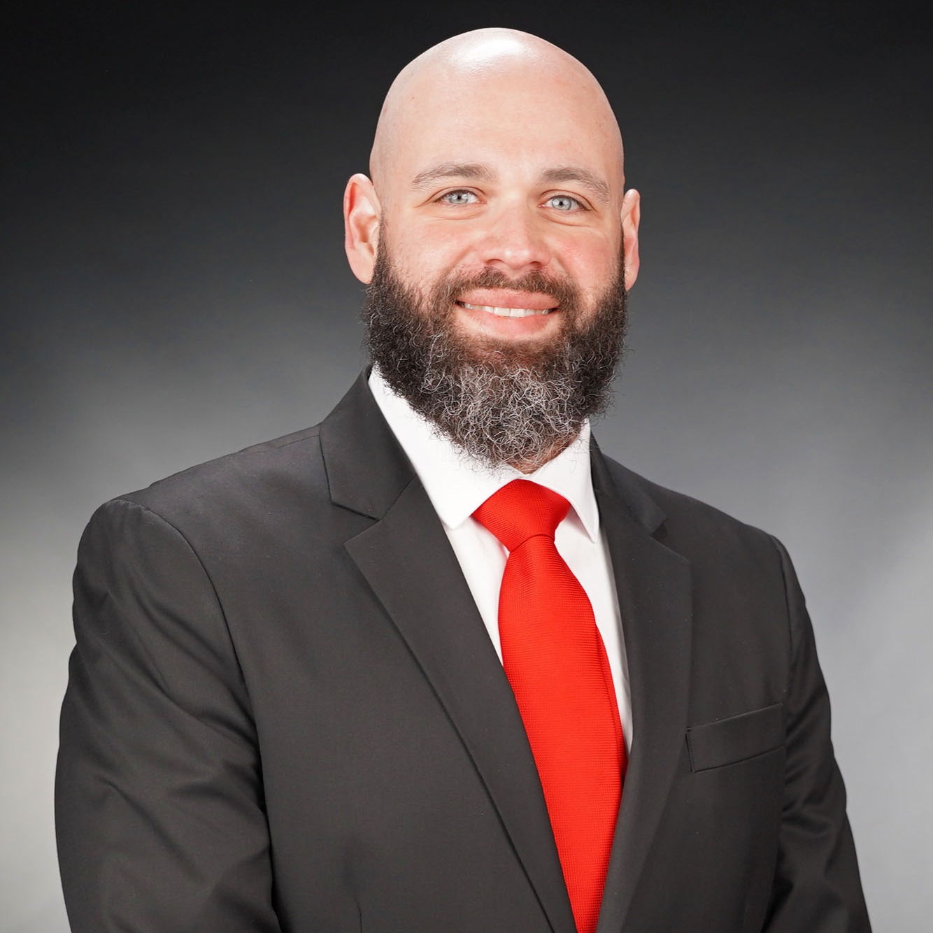 A smiling man with a shaved head and a dark beard wears a black suit and a bright red tie.