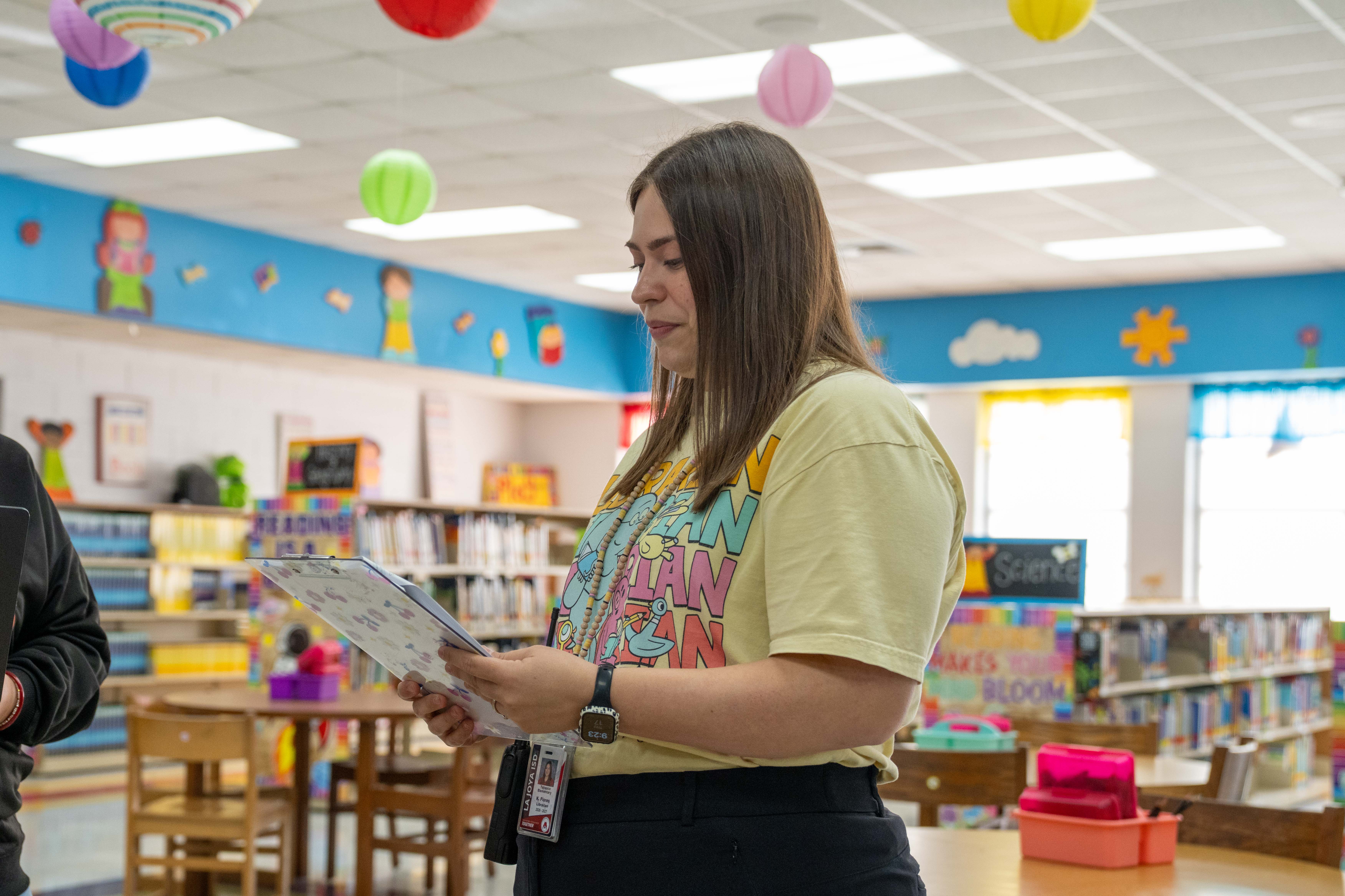 A woman stands in a brightly decorated library, holding and reading from a clipboard.