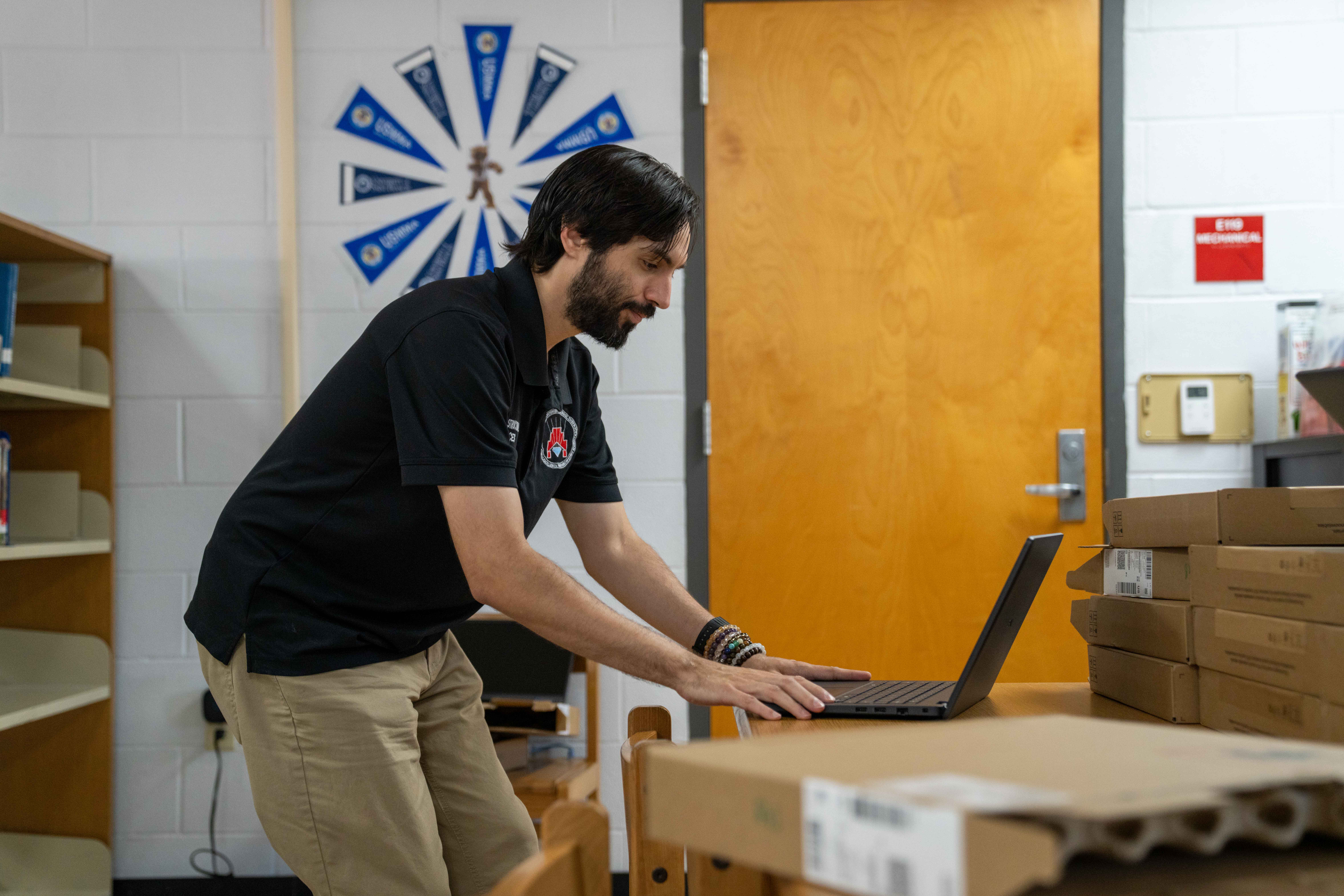 A man in a black polo and tan pants types on a laptop in a room.