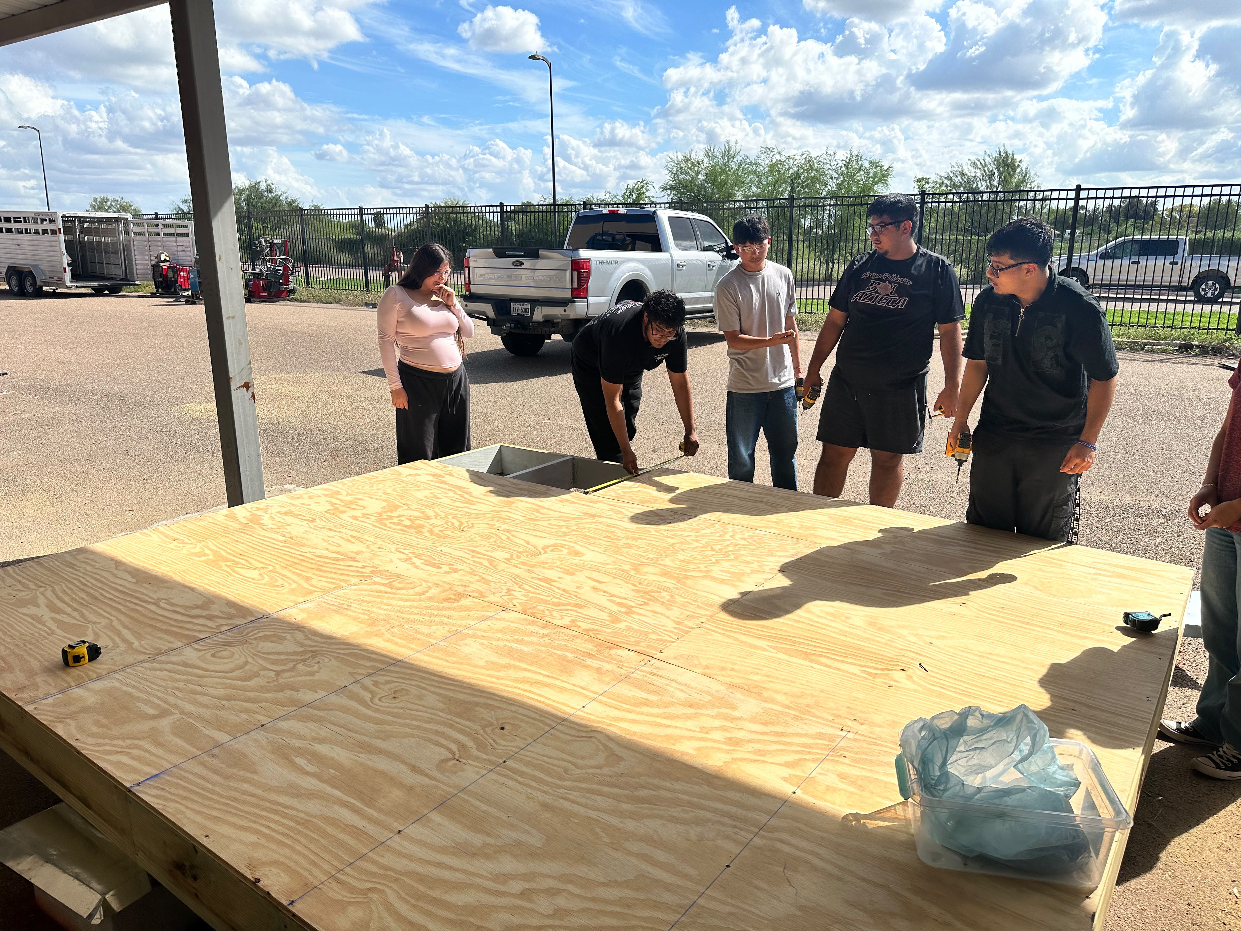 A group of students work together on a large wooden platform outdoors under a partly cloudy sky.