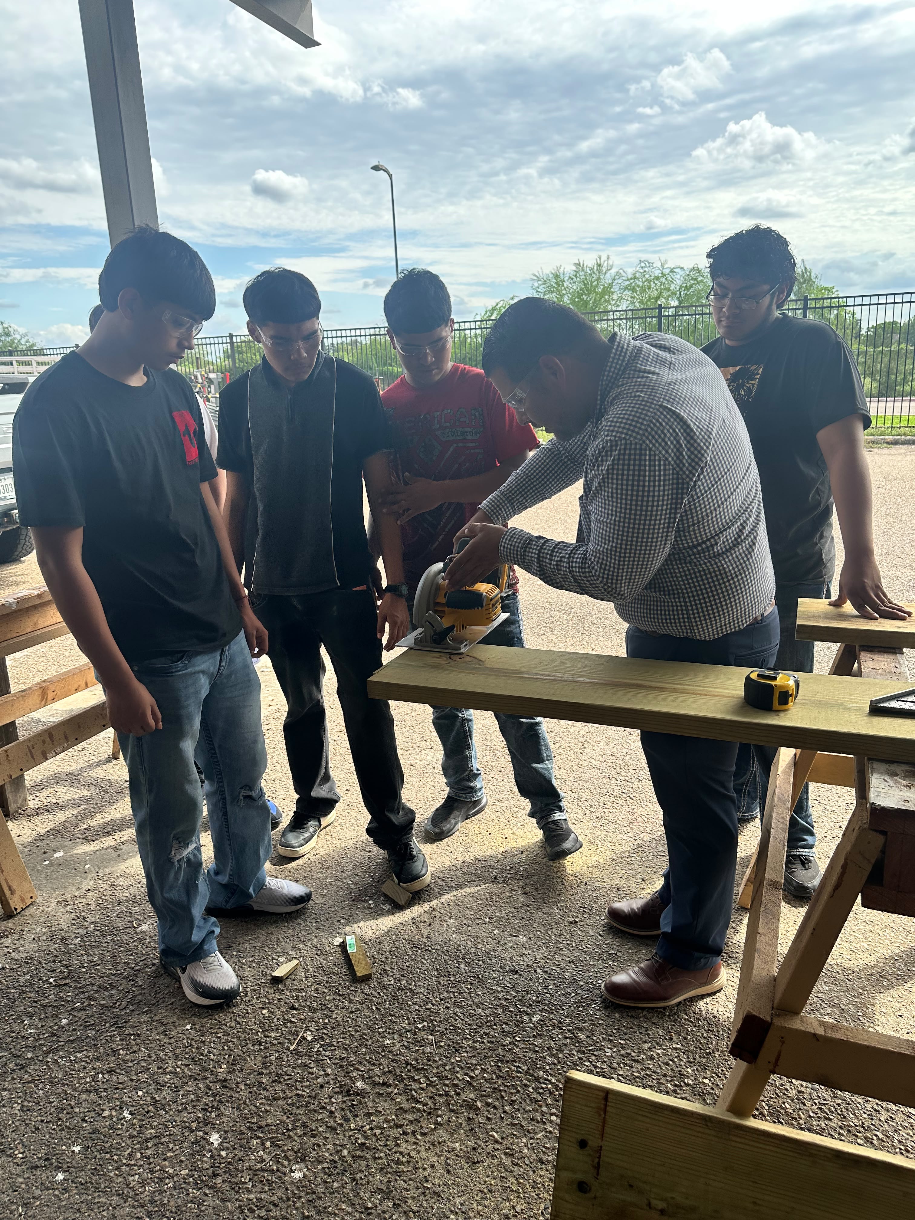 An instructor demonstrates how to use a circular saw on a wooden plank to four students outdoors.