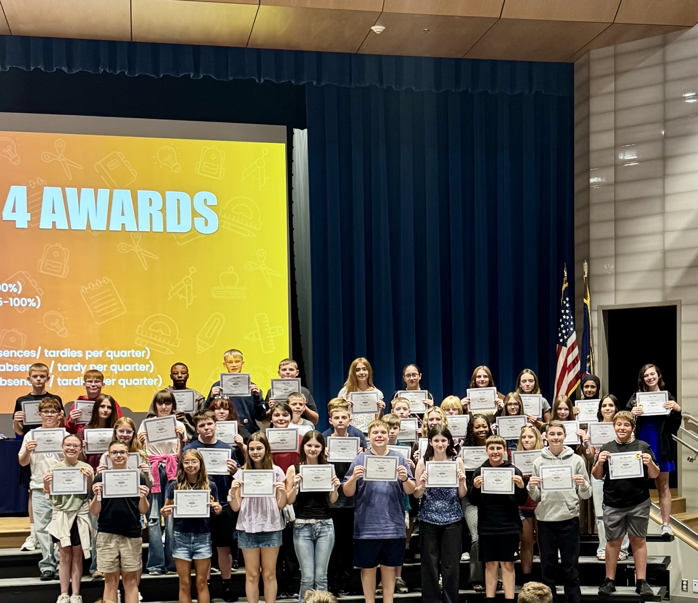 A group of students proudly pose on a stage holding award certificates during a school assembly.