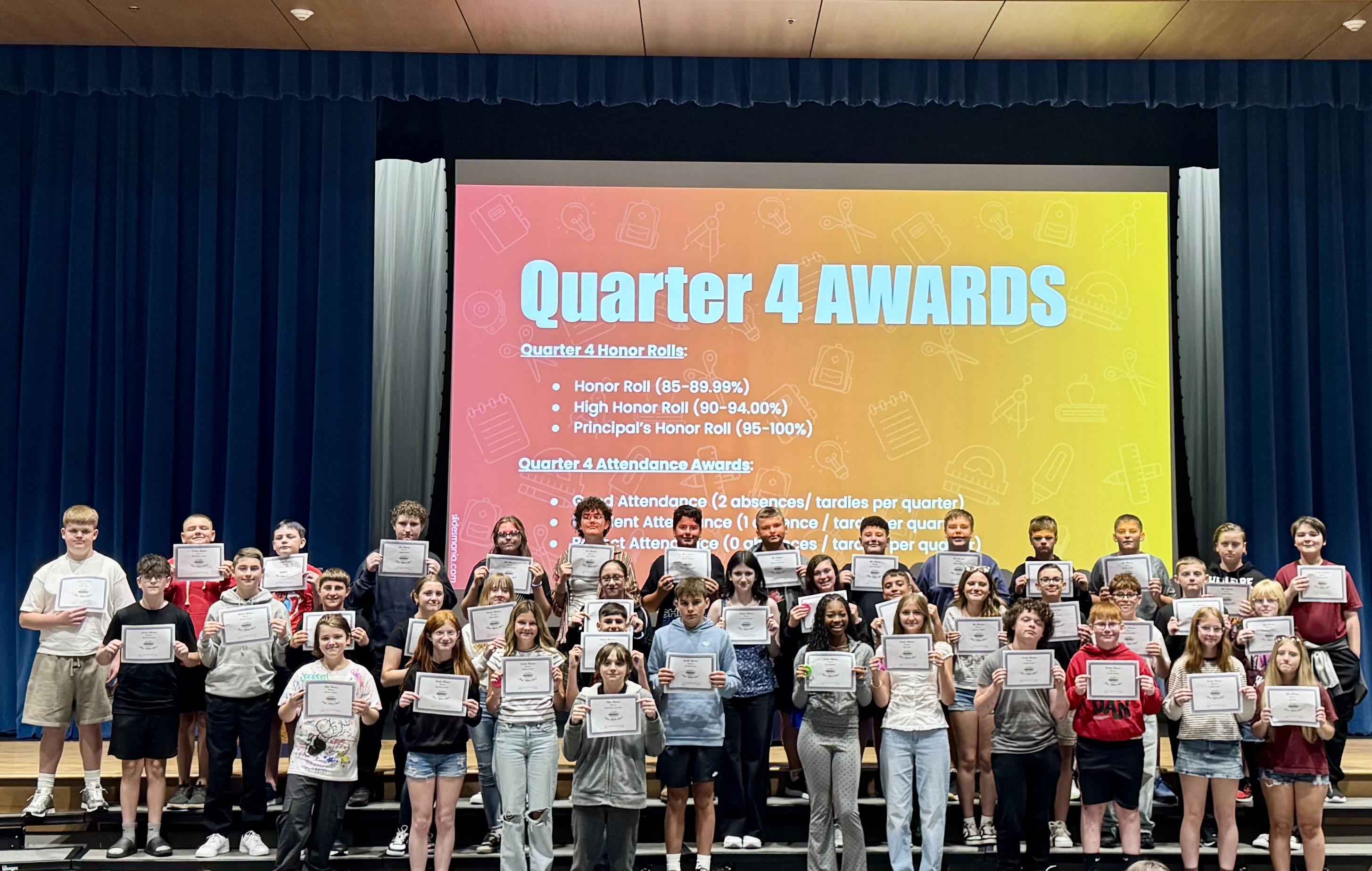 A group of students stands proudly on a stage, holding certificates in front of a large screen that reads "Quarter 4 AWARDS."