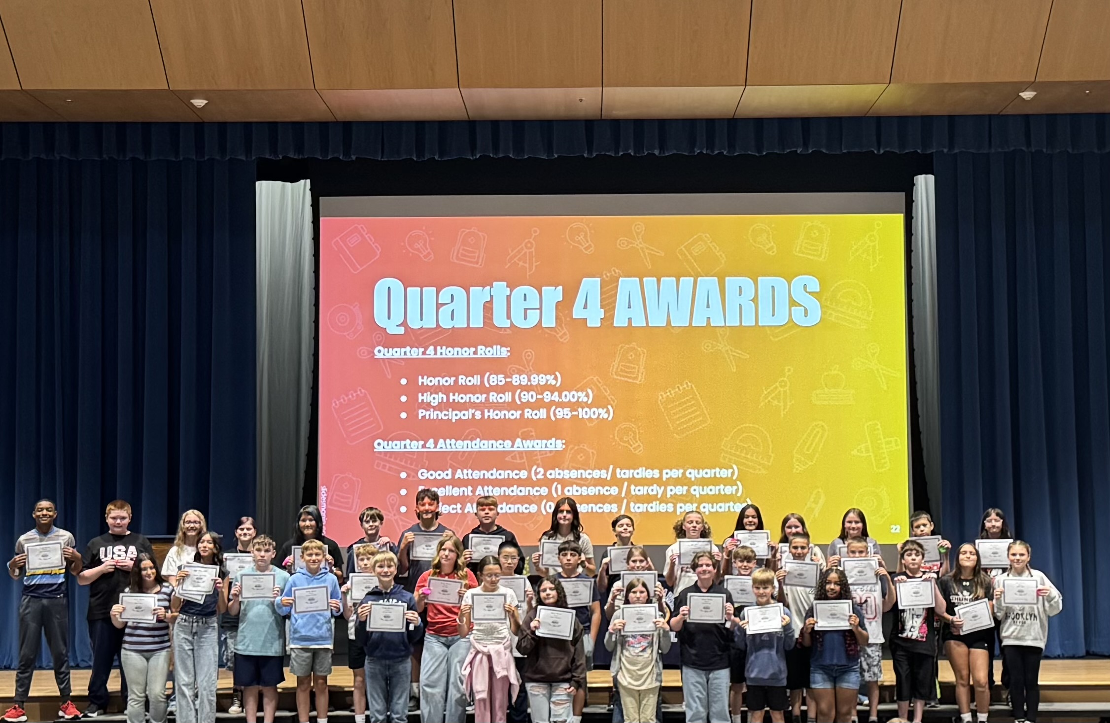 A large group of students proudly hold their certificates in front of a stage screen displaying "Quarter 4 AWARDS."