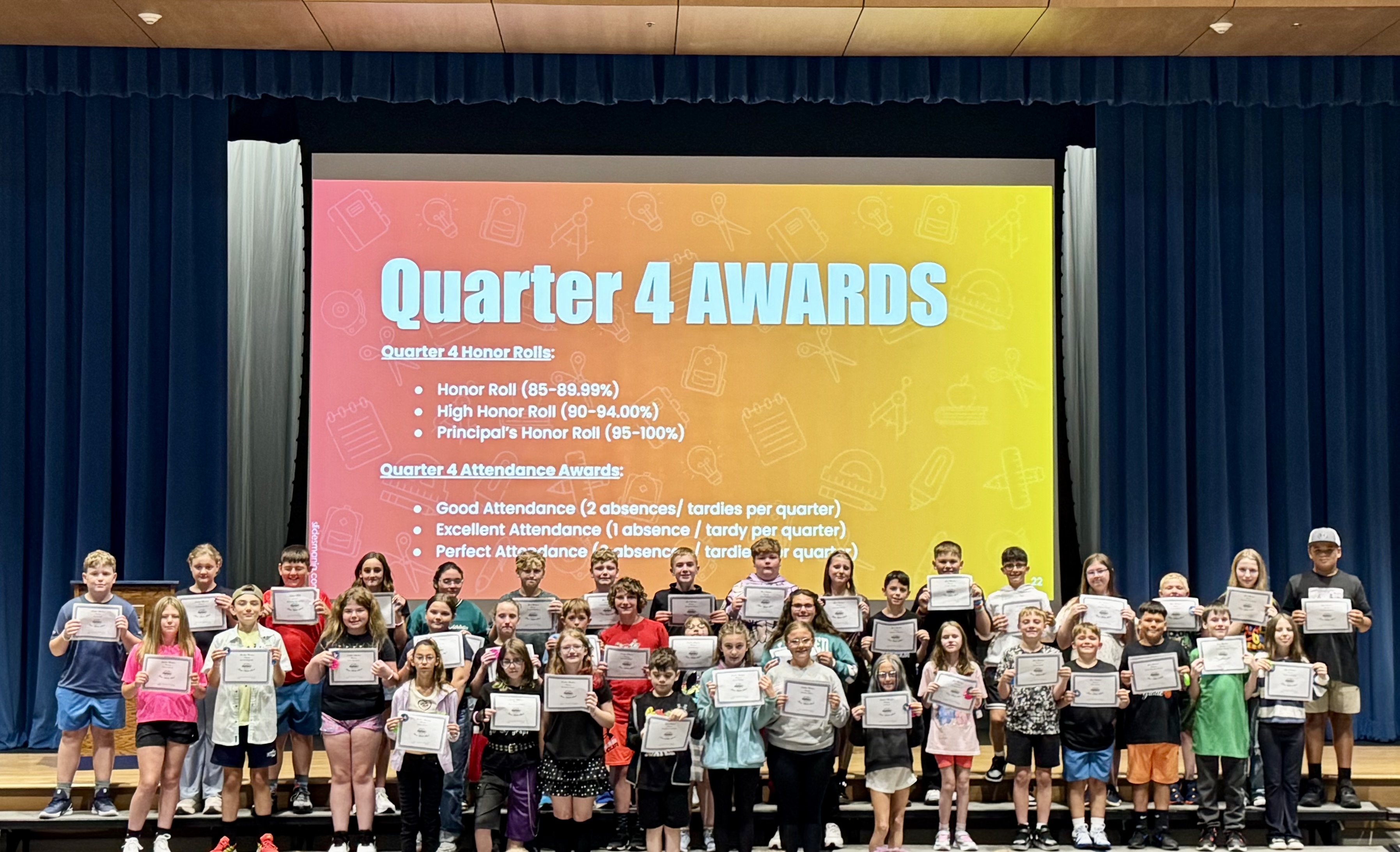 A group of students proudly hold their Quarter 4 Awards certificates on a stage in front of a colorful presentation screen.