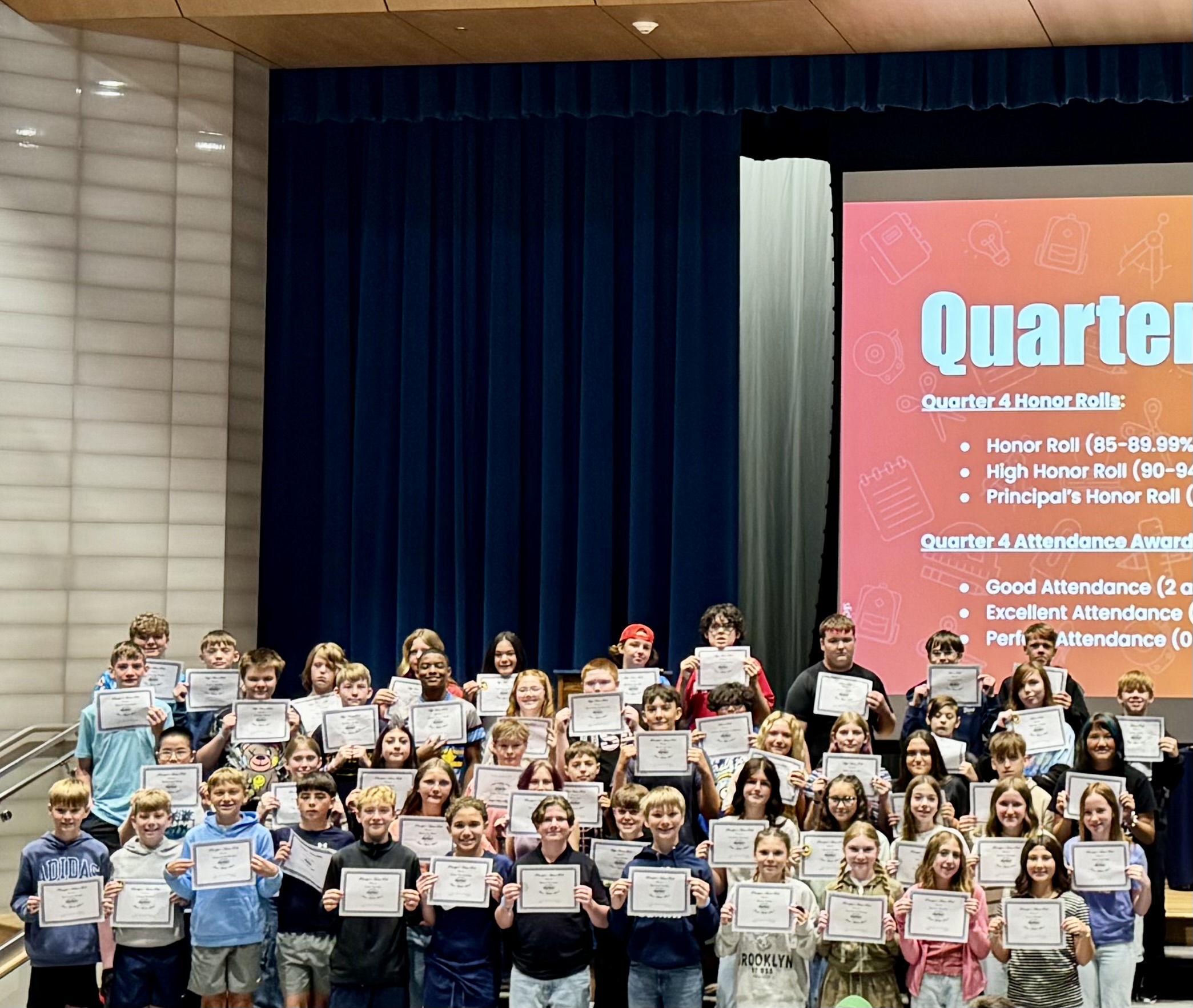 A large group of middle school students stand proudly on a stage holding award certificates during an assembly.