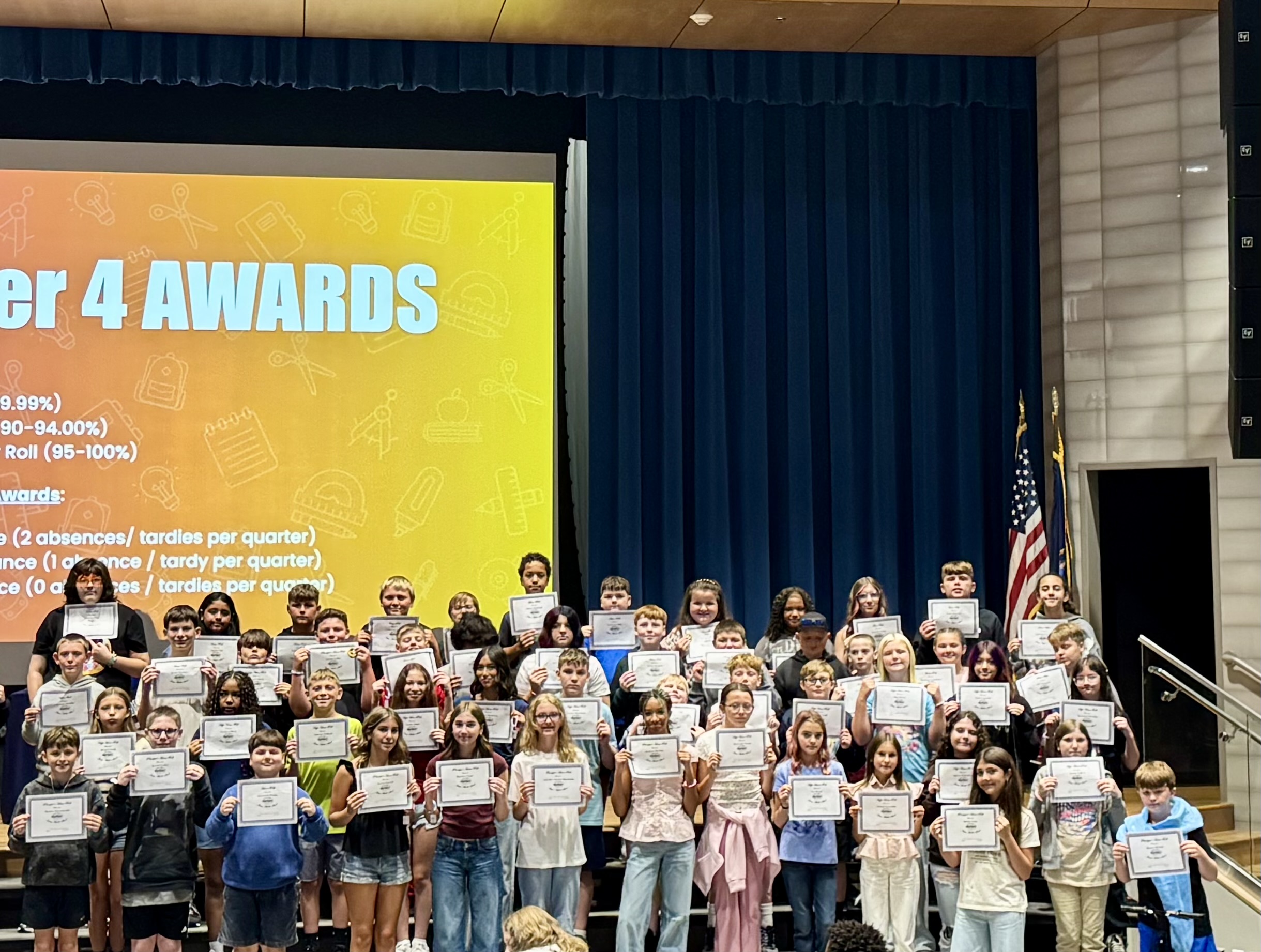 A large group of students proudly hold up their certificates on a school stage during a Quarter 4 Awards assembly.