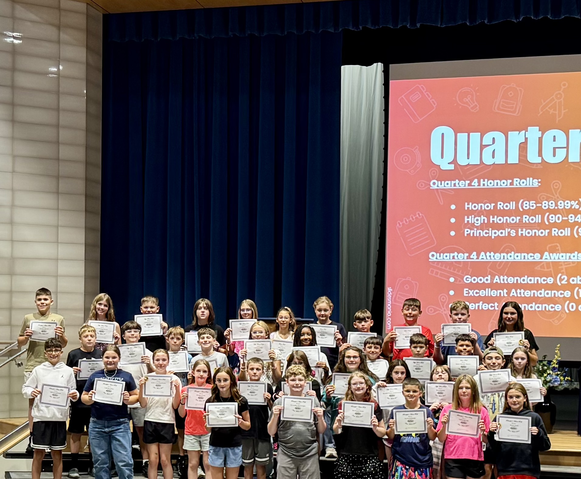 A large group of students proudly hold up their achievement certificates on a stage with a blue curtain backdrop.