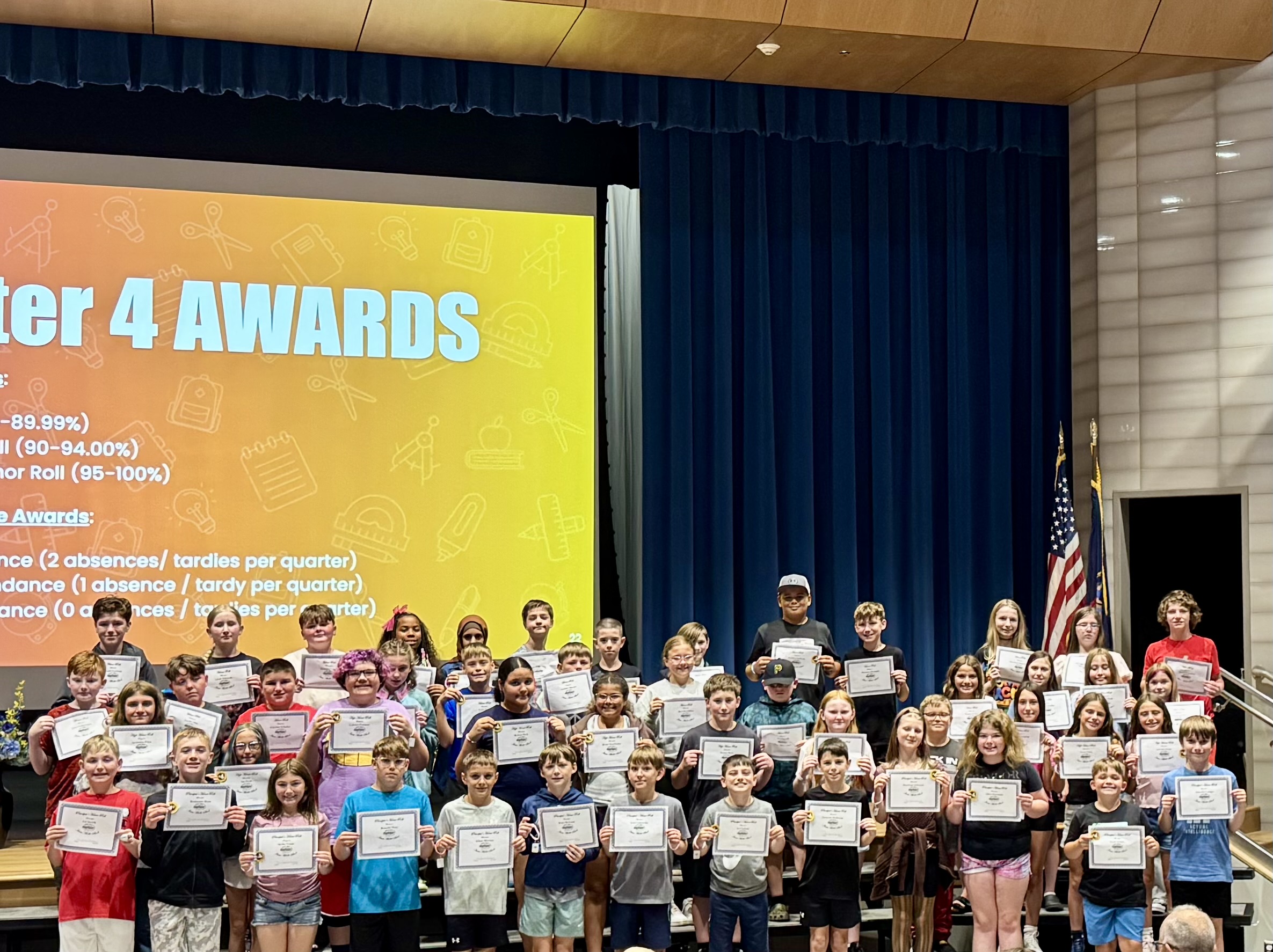 A large group of diverse elementary students proudly hold up their achievement certificates on a school stage.