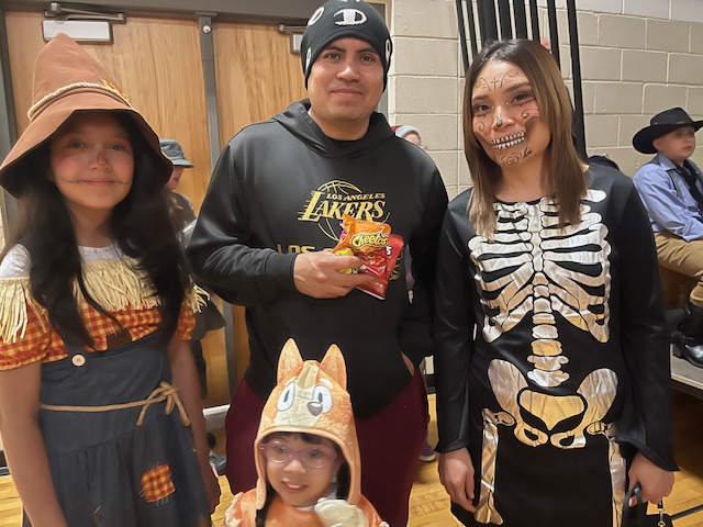 A family poses together in costumes, with one child dressed as a scarecrow, another as an animal, and an adult in a skeleton outfit.