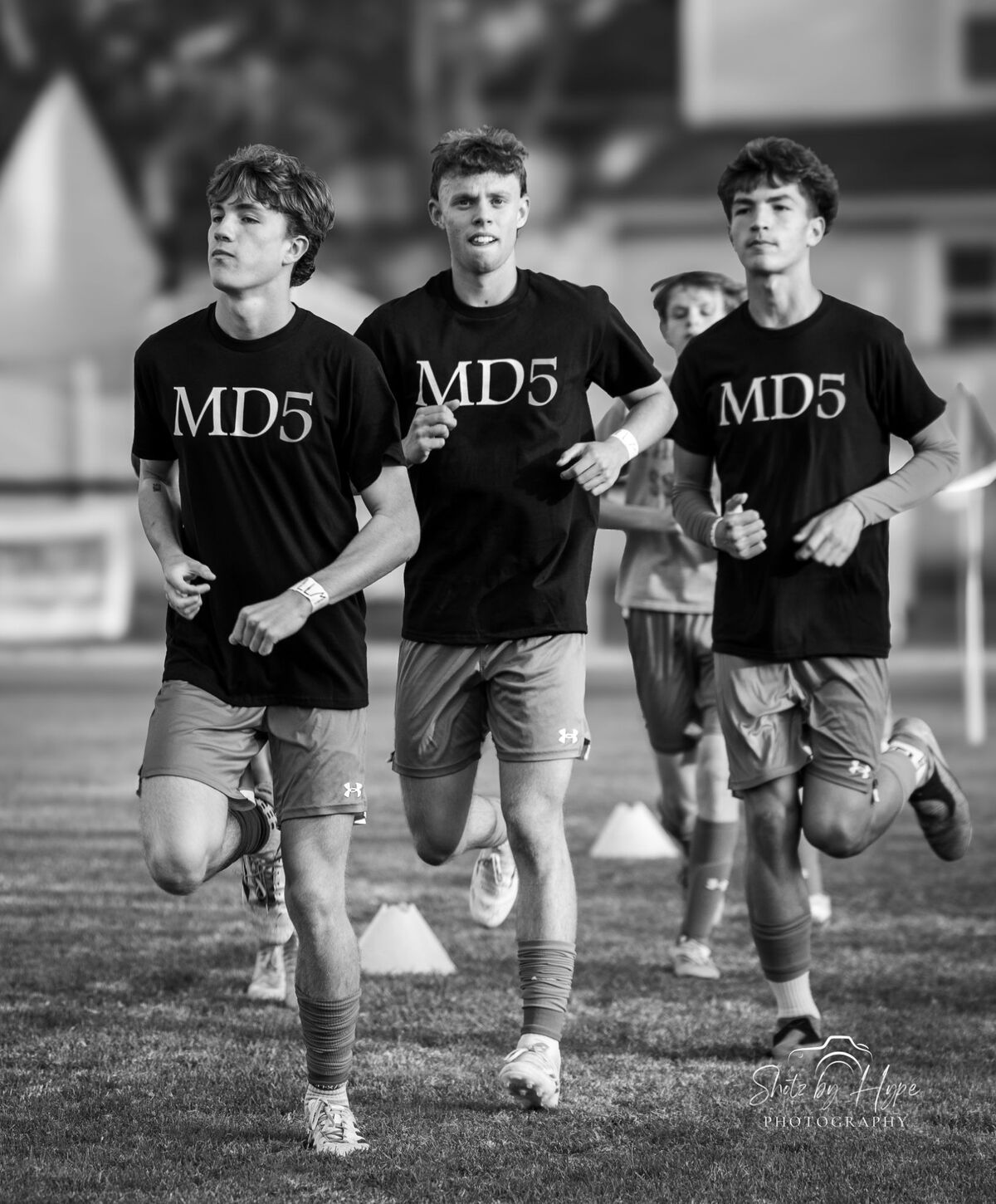 Three young athletes run across a field during a training session, wearing matching black t-shirts with 'MD5' printed on the front.