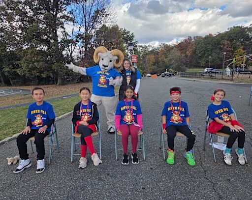 A group of children in blue shirts with 'RIVER HILL' text and medals sit on chairs, with a mascot and adult behind them.