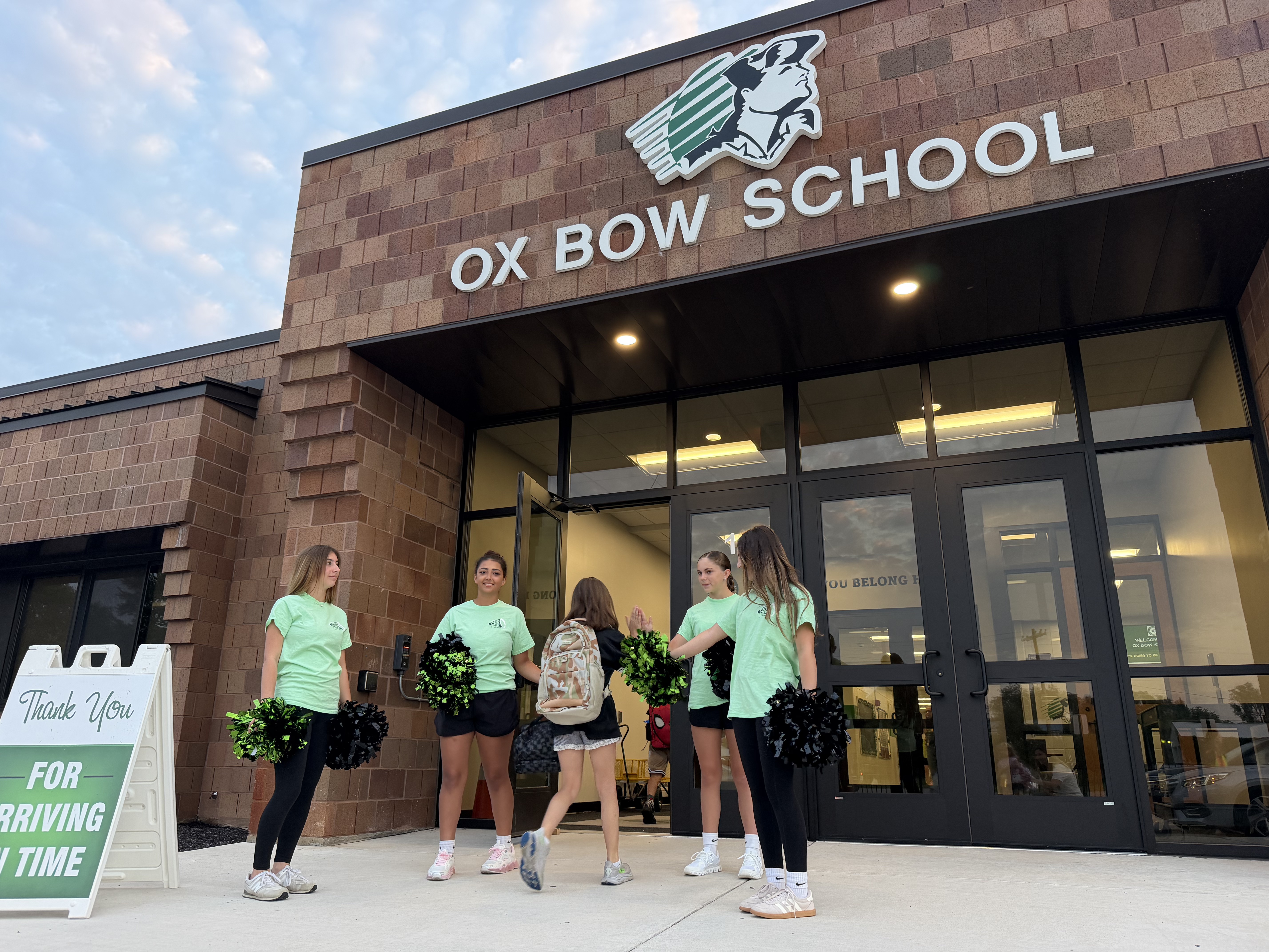 Students in light green shirts and black leggings, holding pom-poms, stand outside Ox Bow School.