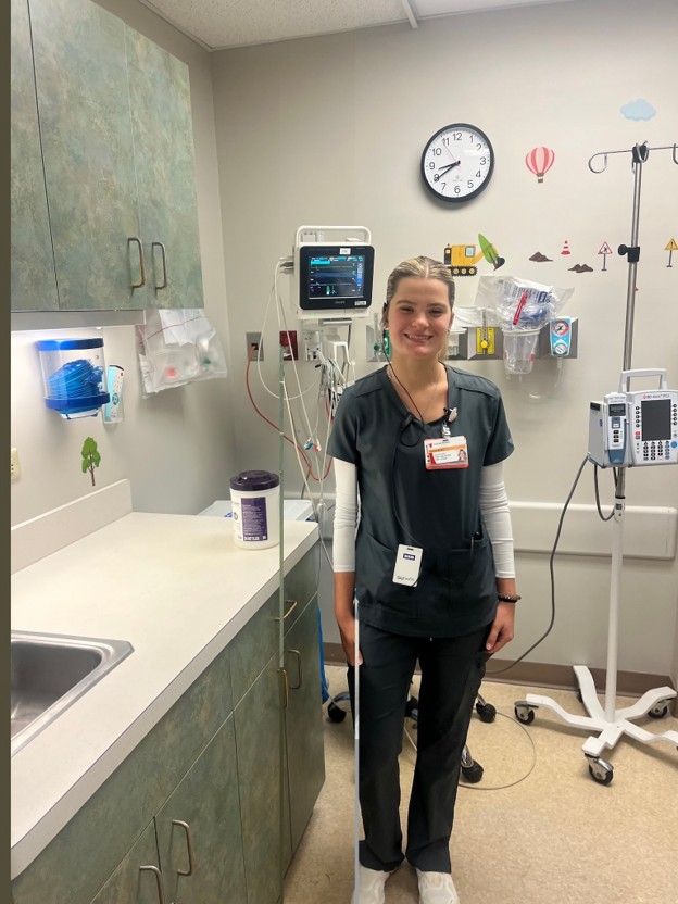 A healthcare worker smiles in a brightly lit pediatric hospital room with medical equipment and wall decals.