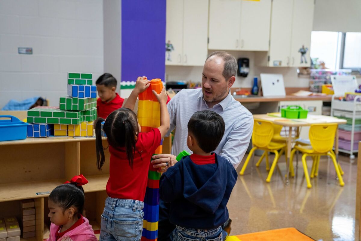 A teacher assists young children in building a tall tower with colorful plastic blocks in a bright classroom.