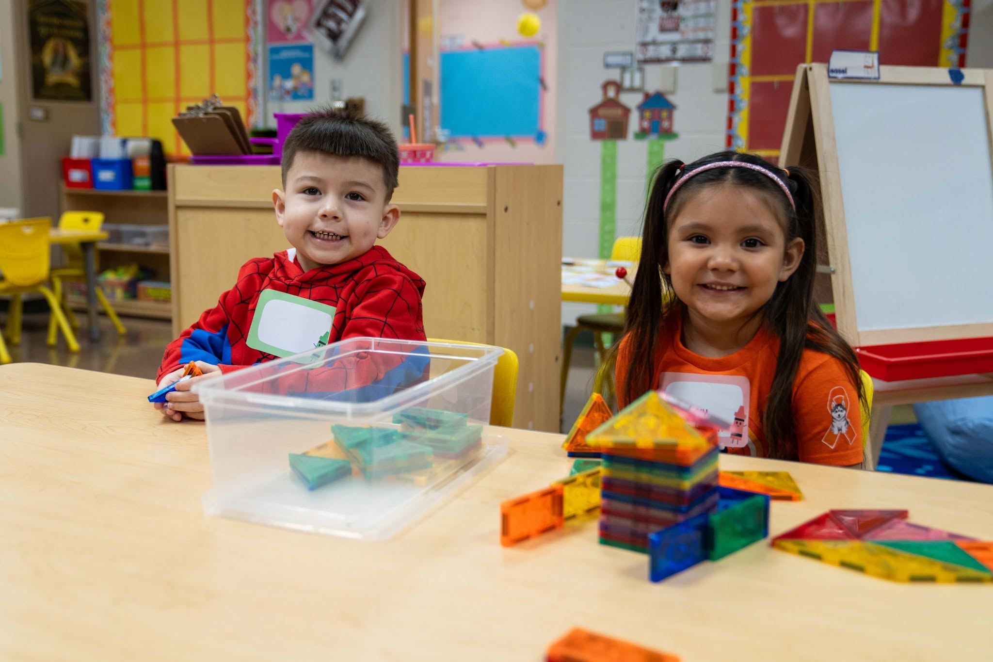 Two smiling children sit at a table in a brightly decorated classroom, playing with colorful magnetic tiles.