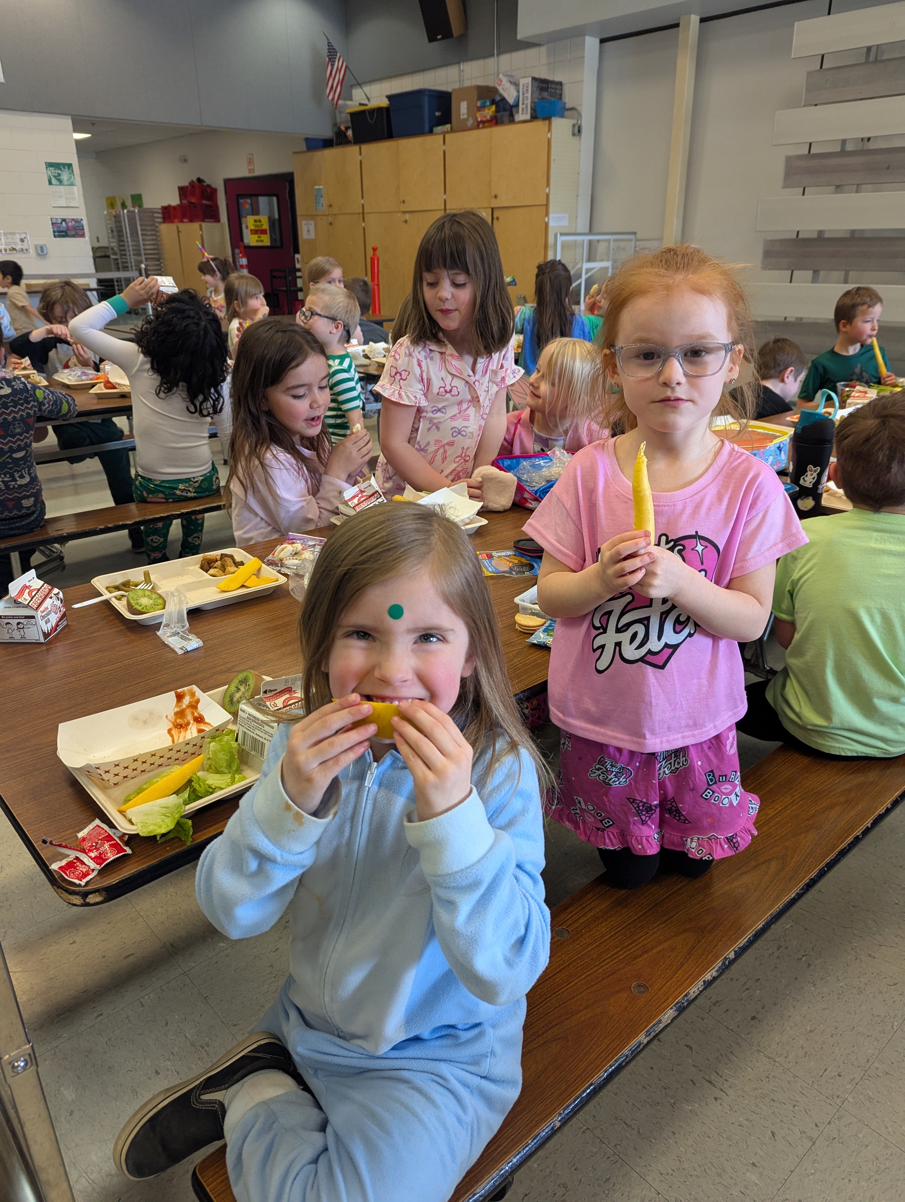Children sit at lunch tables in a school cafeteria, with some eating and others holding food.
