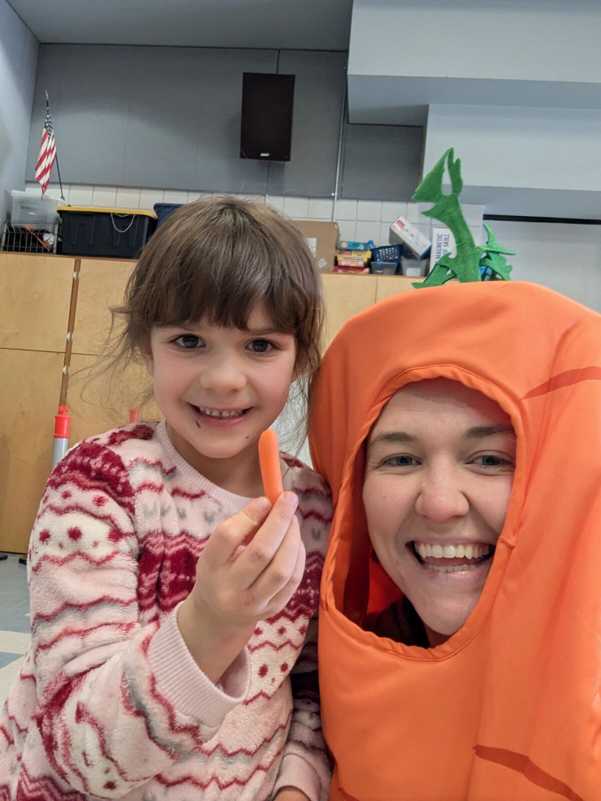 A young girl smiles while holding a baby carrot next to a person in a large carrot costume.