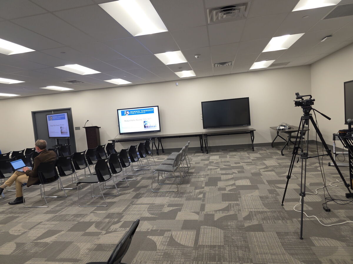 A man sits in a conference room with a laptop, facing rows of empty chairs.